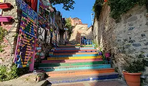 Rainbow-painted stone steps in Balat, Istanbul, surrounded by old stone walls, street art panels, hanging lanterns, and small potted plants.