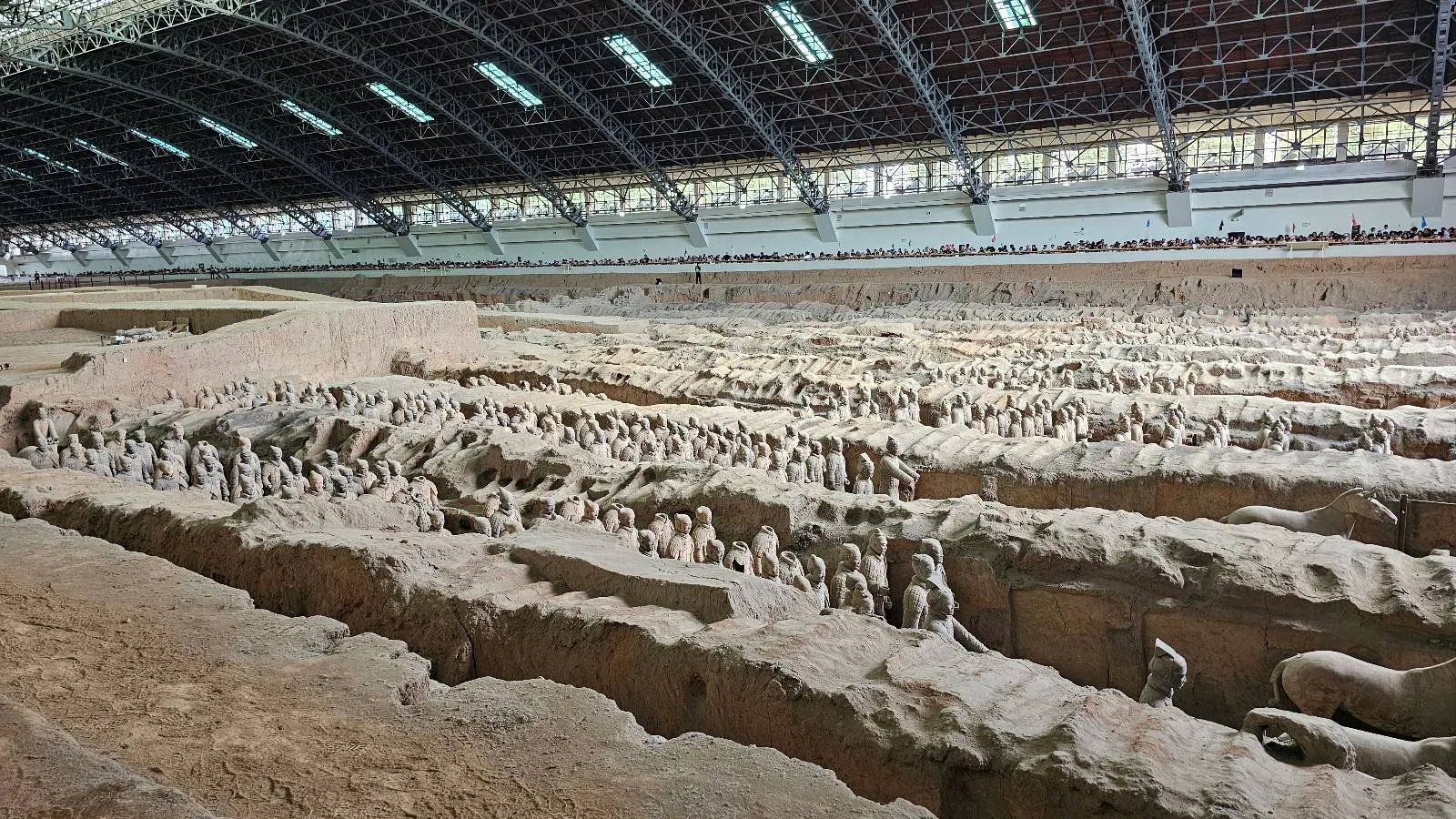 Rows of ancient terracotta warrior statues are partially excavated in a large covered archaeological site, with a high ceiling, natural light, and visitors observing from a raised walkway in the background.