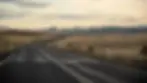 Open road leading to distant hills under a cloudy sky. A car drives along the road with vast grassy fields on both sides and a sign visible.