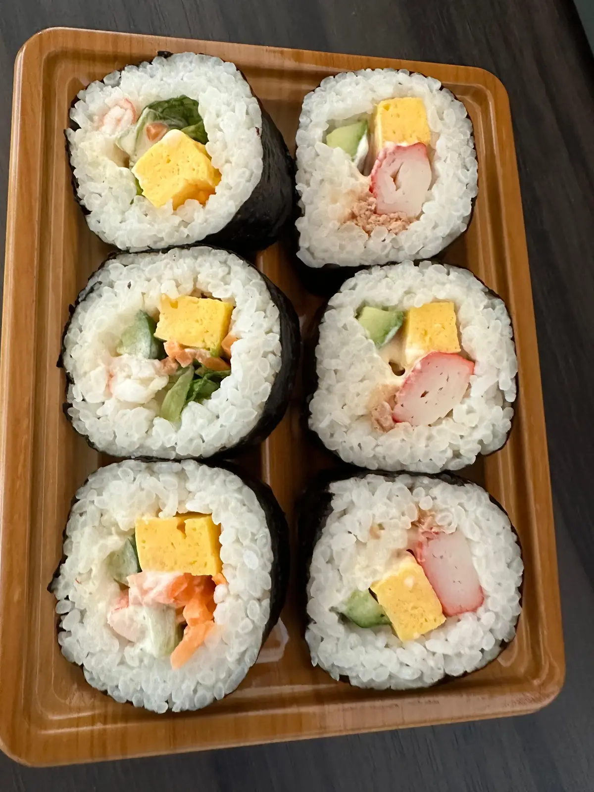 Six pieces of sushi roll on a wooden tray, each filled with rice, imitation crab, cucumber, and a slice of yellow cheese, wrapped in seaweed.