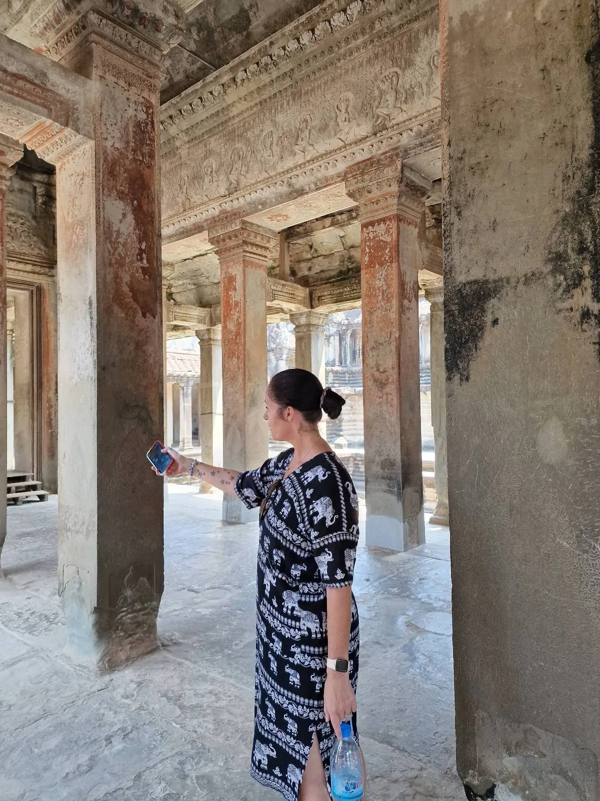 A woman holding a phone stands inside an ancient stone building with tall columns and intricate carvings, looking to her left. Sunlight streams through the open arches, illuminating the architecture.
