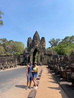 A couple poses in front of an ancient stone gateway with a pointed arch and intricate carvings, embodying the essence of slow travel. Trees flank the entrance as a dirt pathway leads to this hidden gem under a clear blue sky.