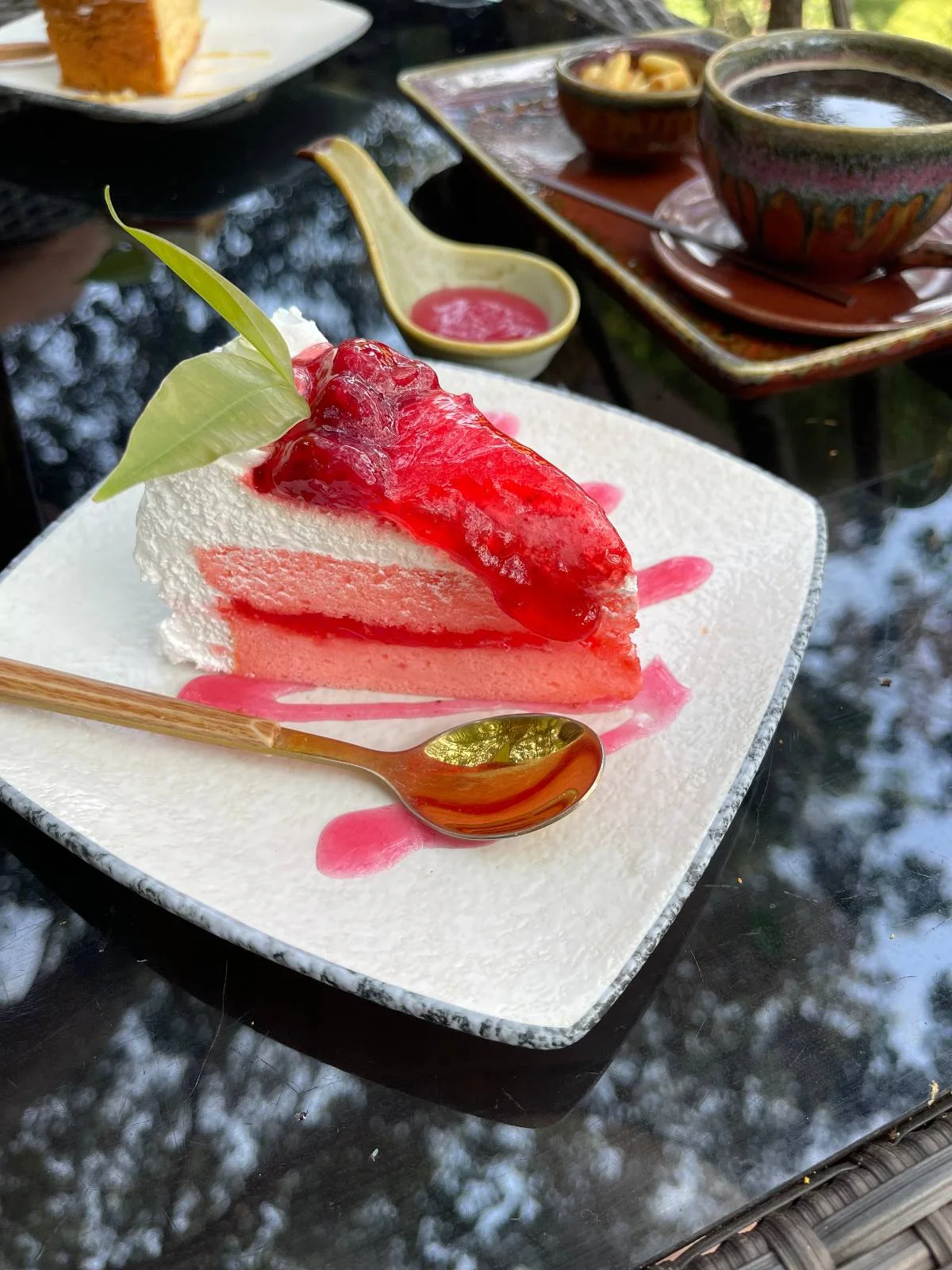 A slice of strawberry cheesecake garnished with a leaf on a white square plate. A wooden spoon with sauce accompanies it. A blurred background shows a table with more dishes and a lush green view.