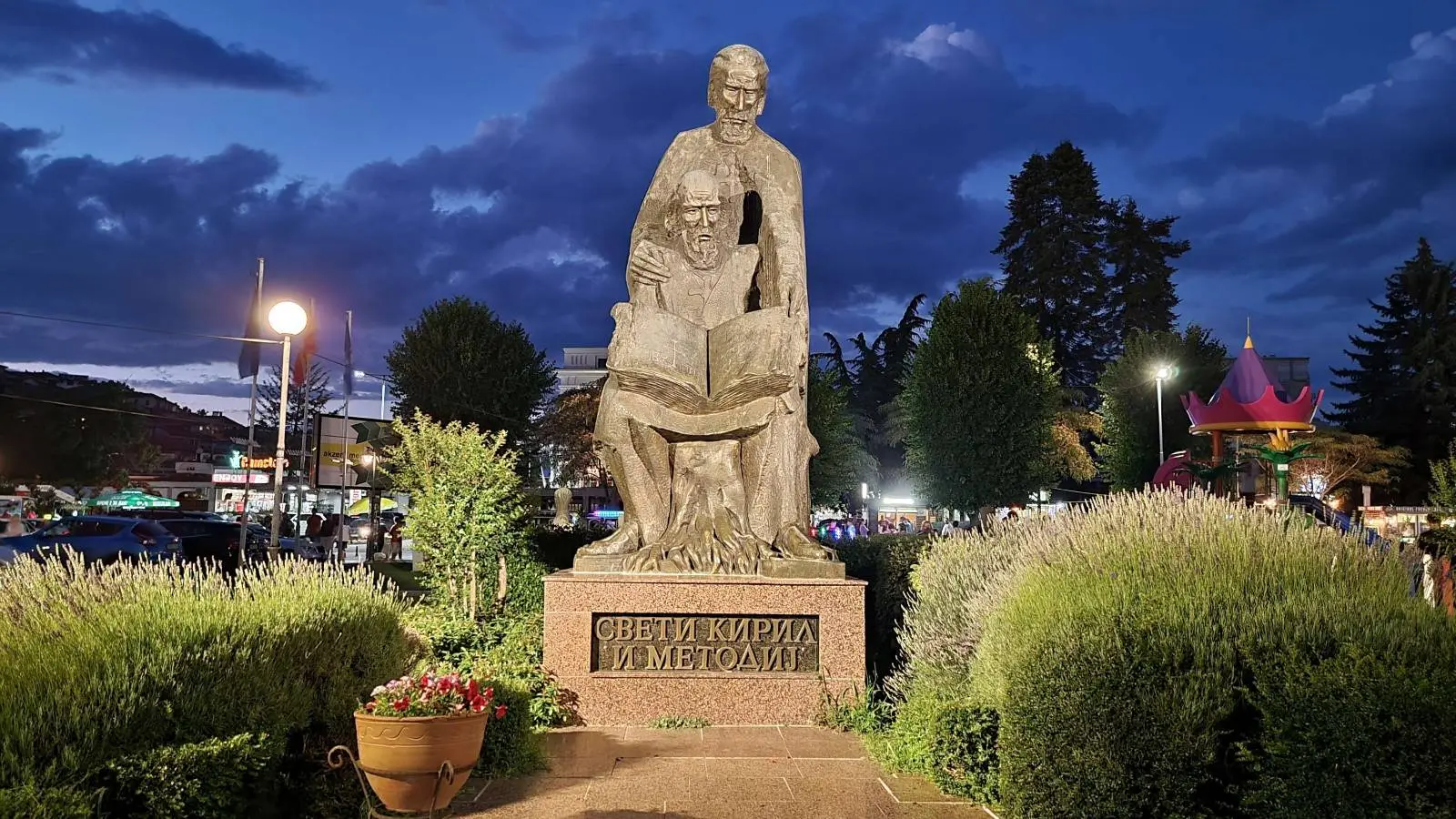 Statue of two figures reading a book at night, surrounded by greenery and colorful lights. Text on the base reads: "Свети Кирил и Методий".