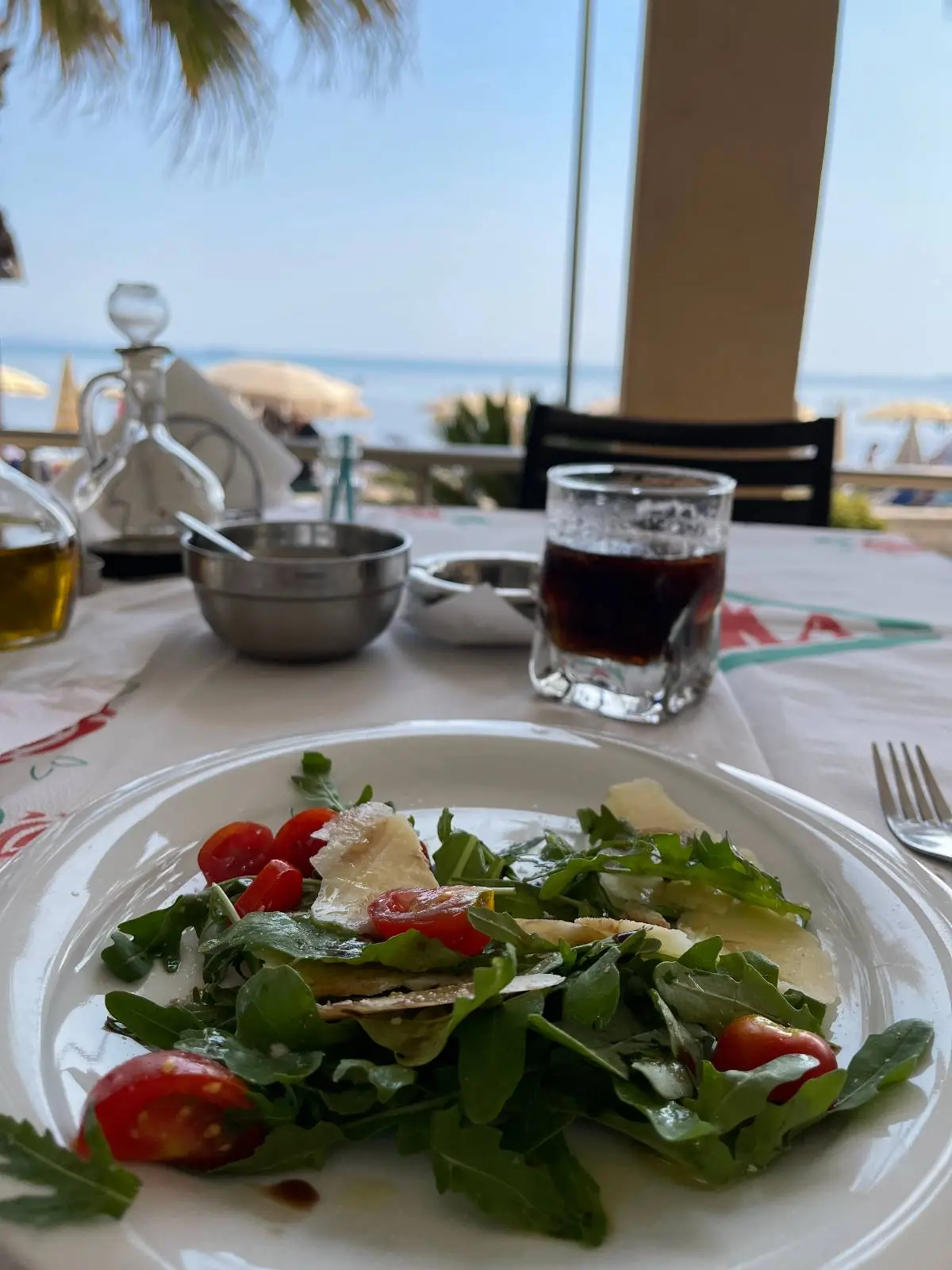 A plate of fresh salad with cherry tomatoes and greens sits on a table, next to a glass of red drink, with a seaside view in the background.