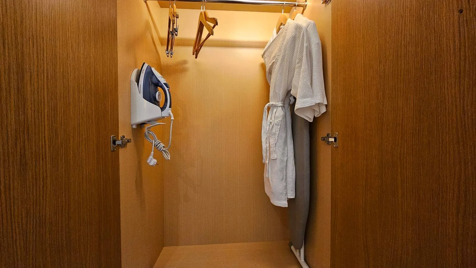 A wooden wardrobe with doors open, showing an iron, ironing board, hangers, a set of towels, and a white bathrobe hanging inside. The wardrobe interior is well-lit.