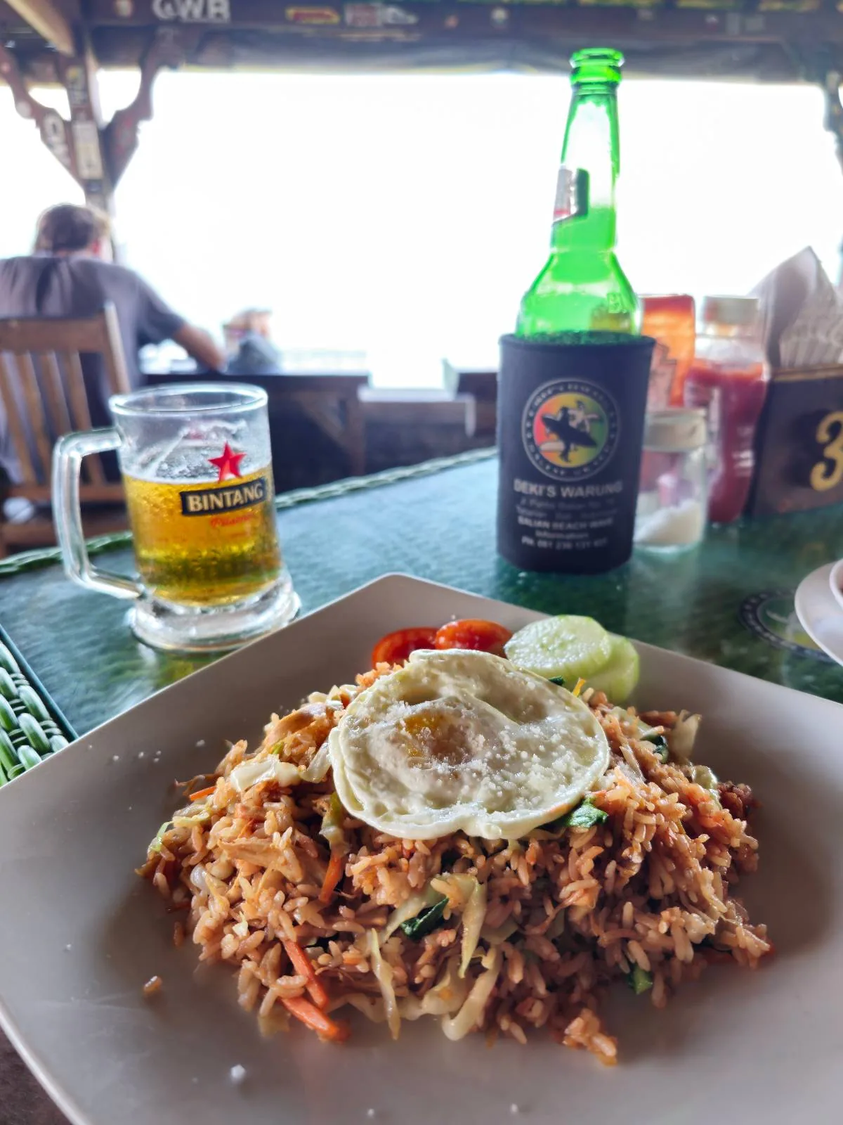A plate of fried rice topped with a fried egg is on a table alongside a glass of Heineken beer and a bottle in a koozie. The background shows a person sitting at a table and a view of the ocean.