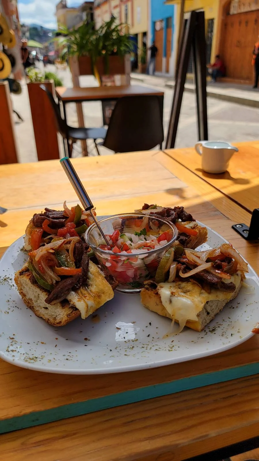 A plate of sliced bread topped with melted cheese, vegetables, and meat surrounds a small dish of tomato-based salsa. The dish is on a wooden table in an outdoor café with a street and buildings visible in the background.