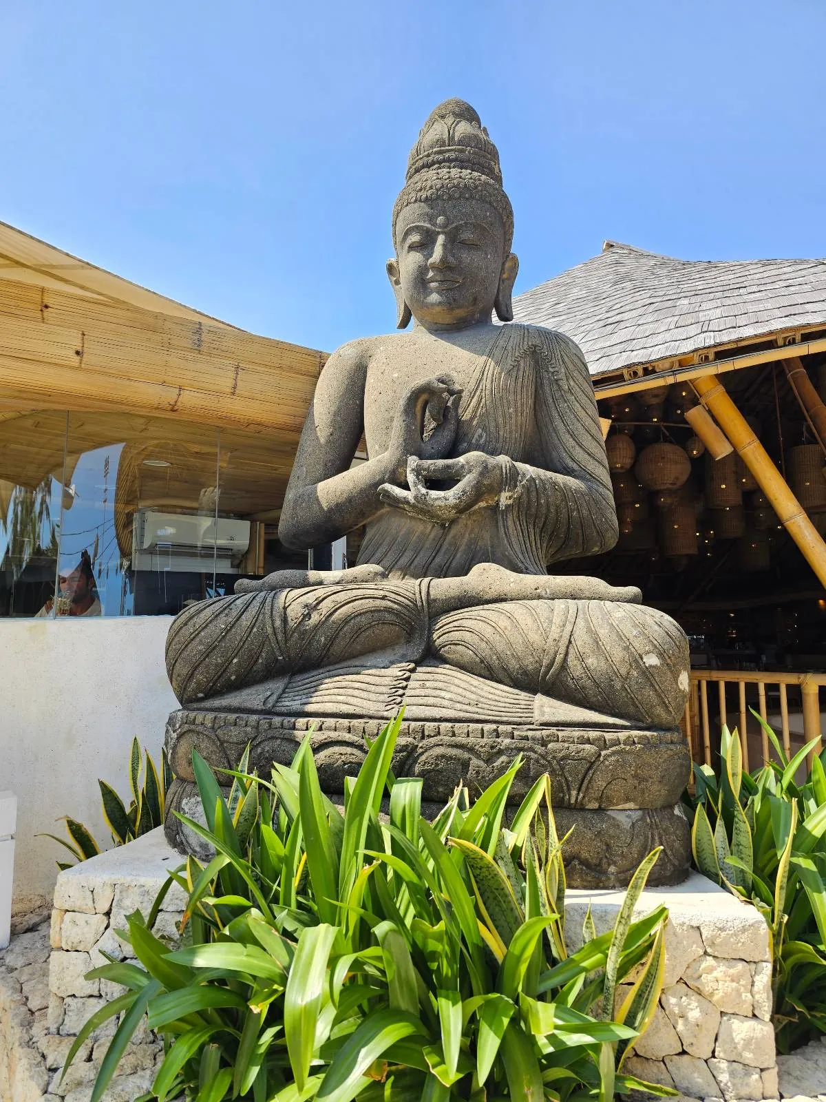 A large stone statue of Buddha sits cross-legged on a pedestal surrounded by green plants. The Buddha is in a meditation pose with hands in a mudra. A thatched roof structure is partially visible in the background under a clear blue sky.