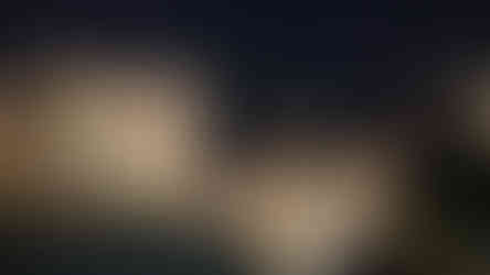 Night view of a white building with balconies and lit windows, overlooking a calm body of water with reflections and distant lights in the background.