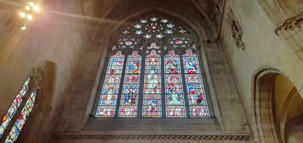 Stained glass window in a cathedral interior, depicting vivid biblical scenes in red and blue hues. Sunlight beams through, creating a serene atmosphere.