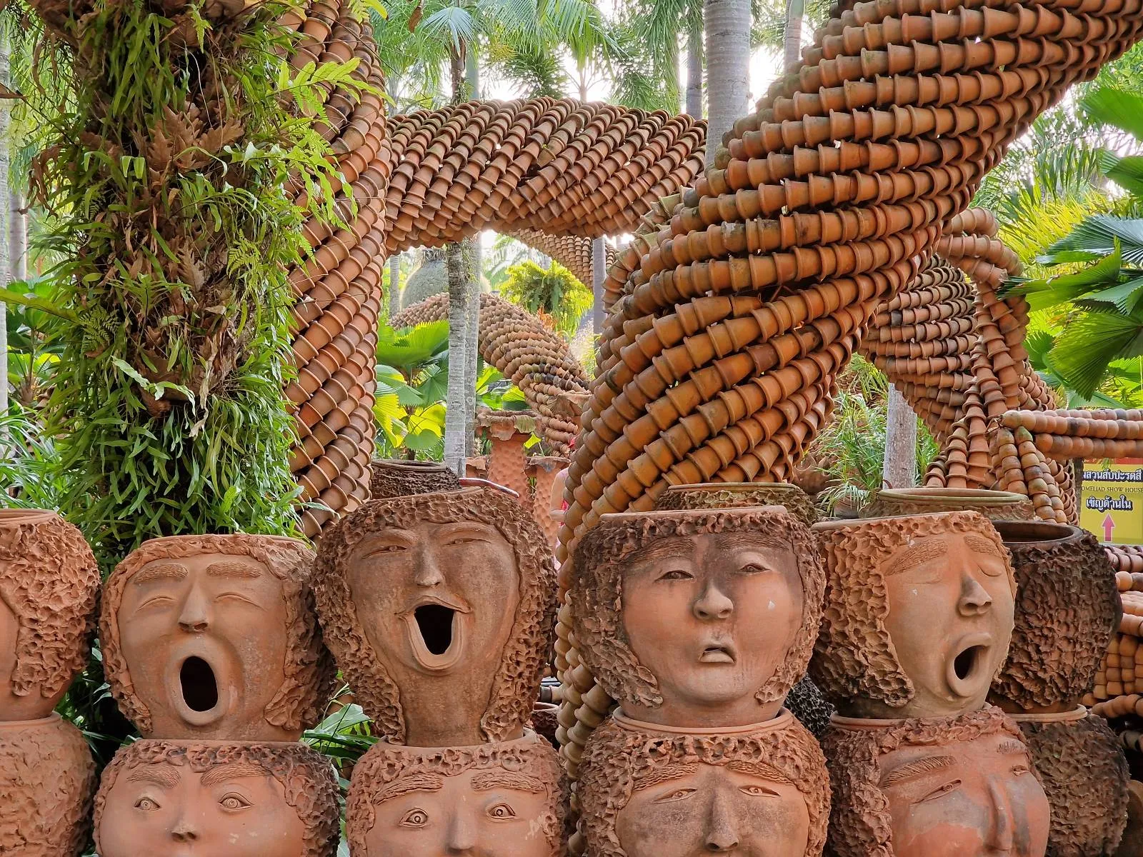 A collection of clay pots shaped like expressive human faces, some with open mouths, are on display in a garden. They are arranged in front of a lush background, featuring palm trees and large spiral woven structures. The atmosphere is vibrant and whimsical.