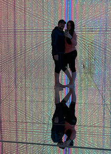A couple takes a selfie in a room filled with colorful, reflective LED lights. The exhibition's walls and floor create an immersive, mirrored effect, surrounding them in vibrant hues that offer visual stimulation.