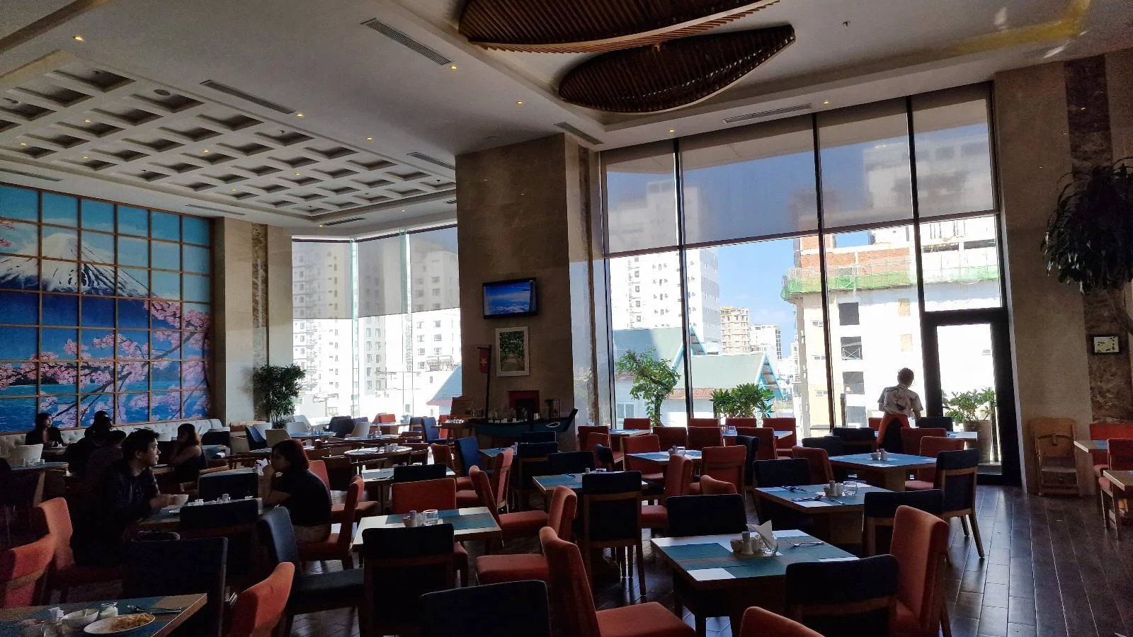 A spacious, modern restaurant with high ceilings and large windows allowing natural light to fill the room. Tables and chairs are neatly arranged, and a wall features a colorful mural. Some people are seated, enjoying their meals.