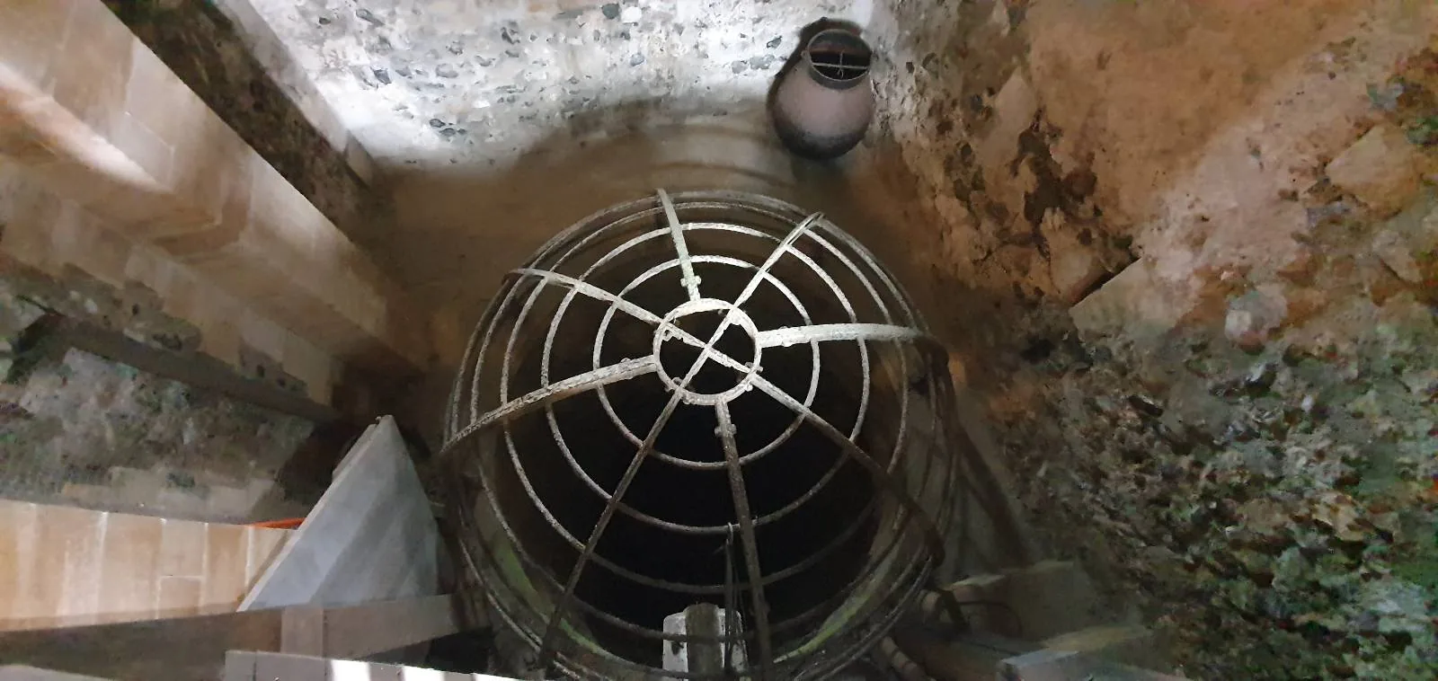 View down a narrow, cylindrical stone well with a metal grille covering the opening. The walls are aged and textured, and there's a round, clay pot or jar near the top, resting on the edge.