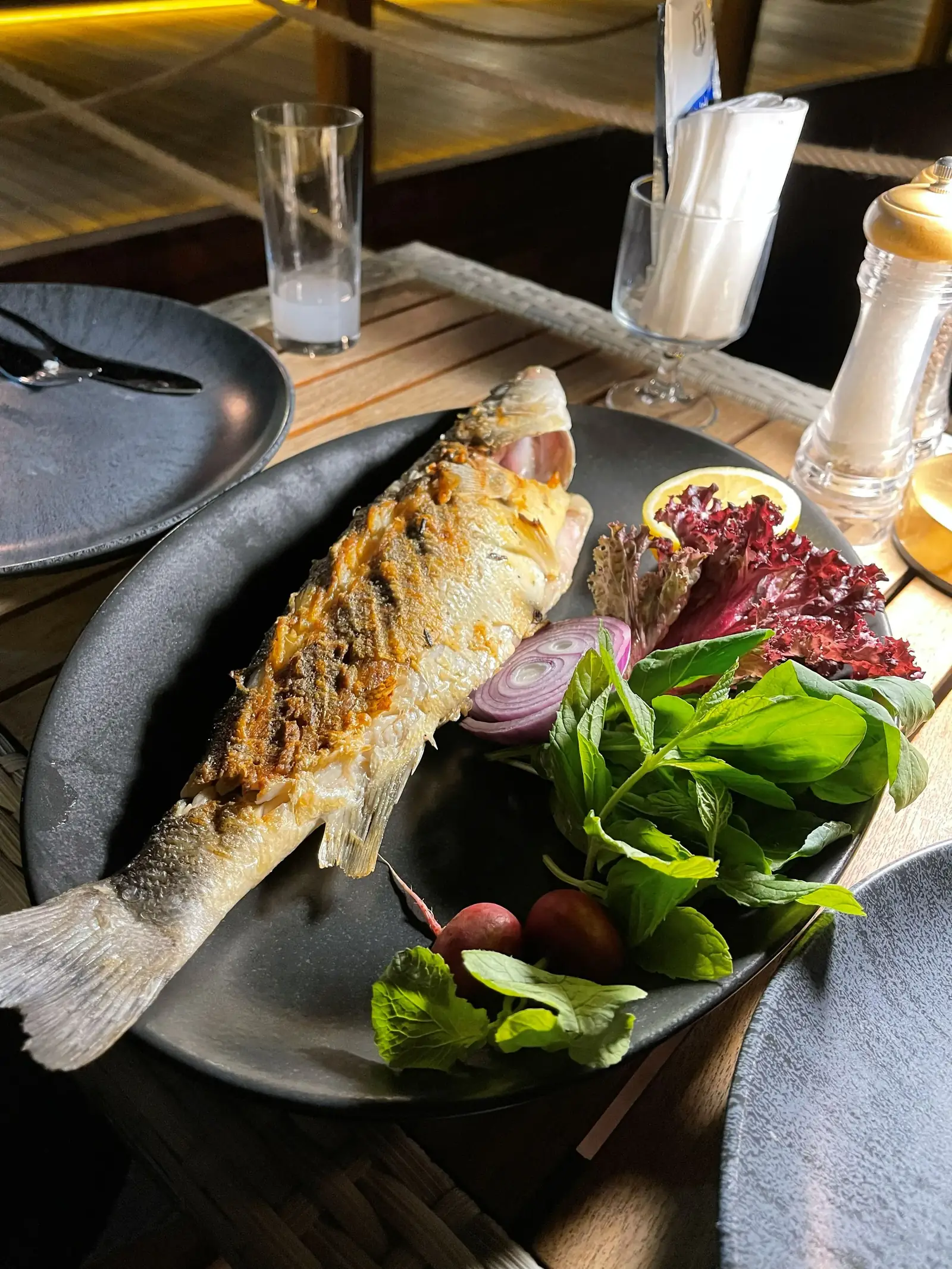 Grilled whole fish on a black plate, garnished with lemon, lettuce, purple onion slices, and radishes. A glass, utensils, and napkins are in the background.