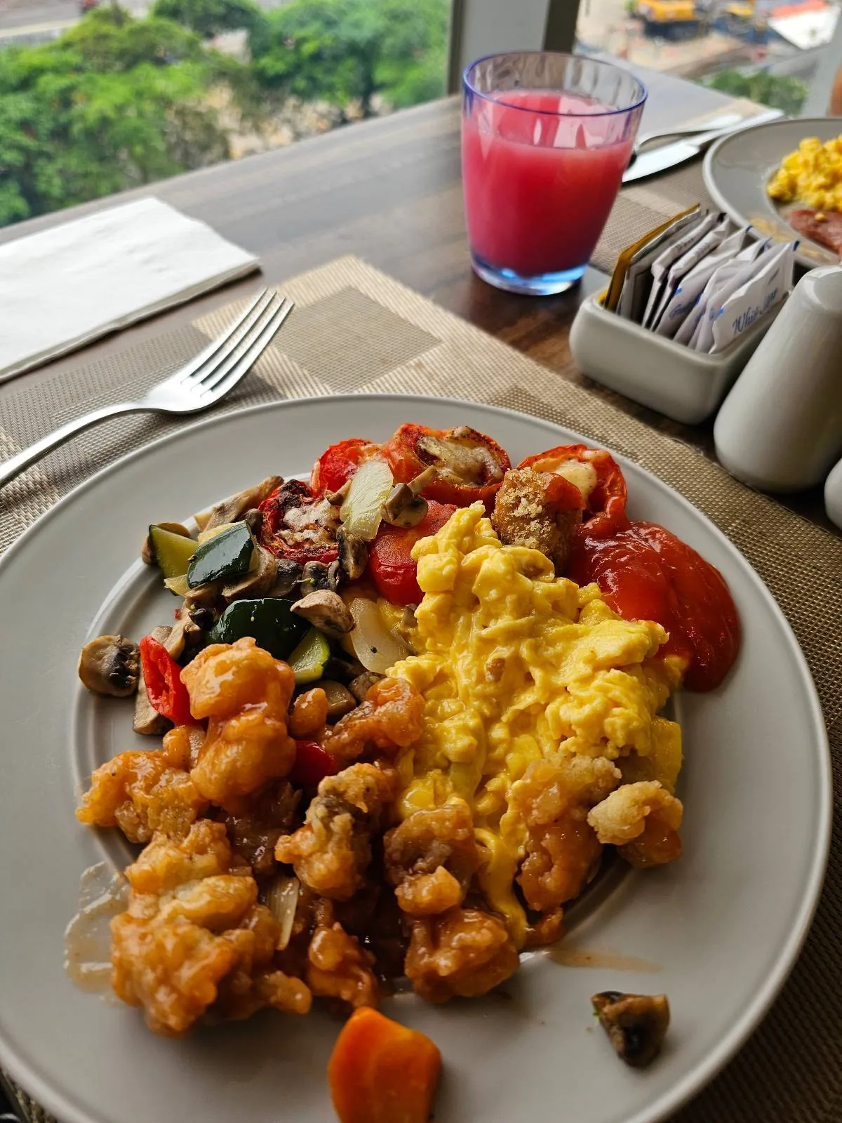 A plate of scrambled eggs, fried chicken pieces, and roasted vegetables like zucchini and tomatoes, set on a table near a window. A glass of pink juice and a container with sugar packets are in the background.