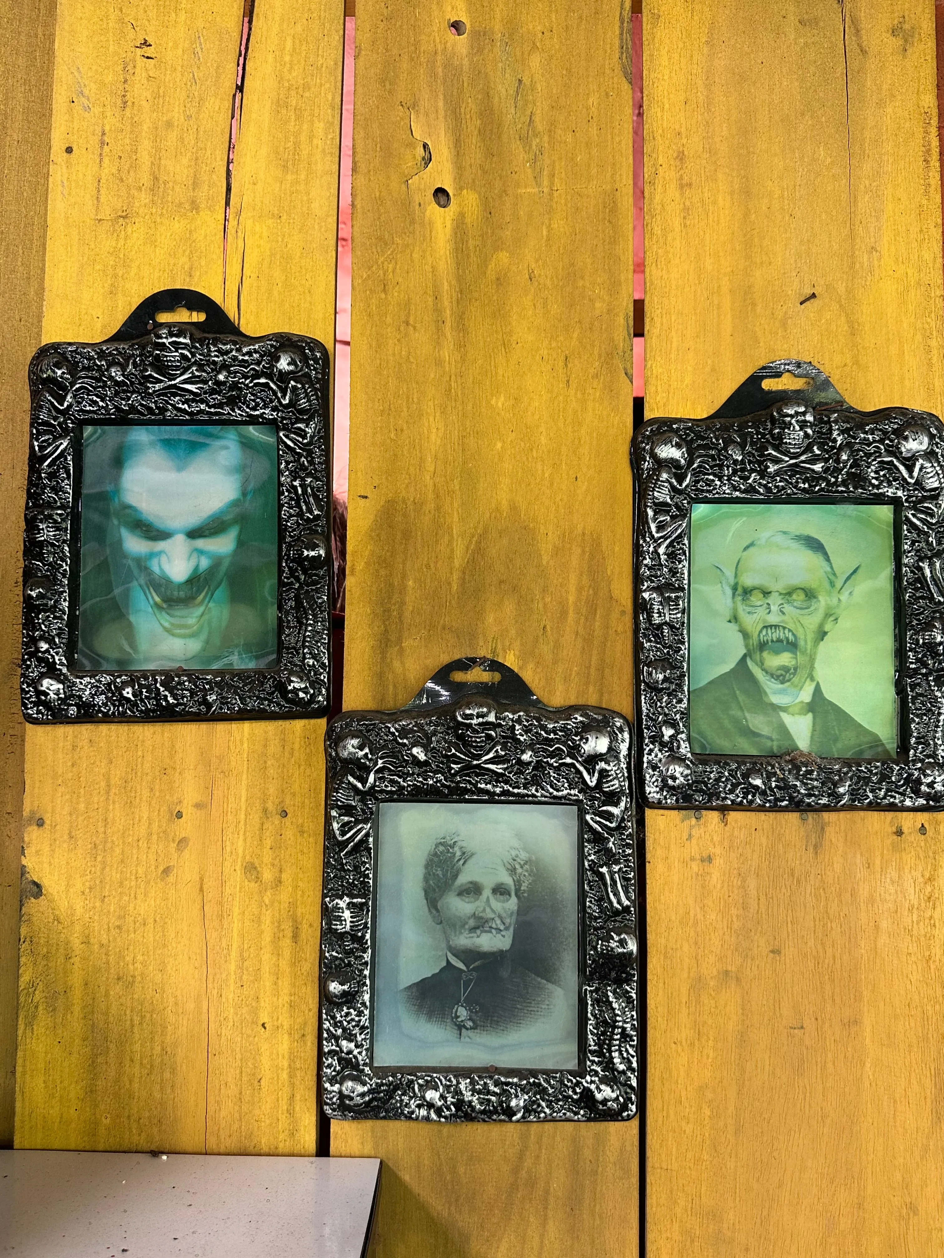 Three ornate, black-framed vintage portraits hang on a yellow wooden wall. The top left portrait appears ghostly or distorted, while the other two show an older man and woman in old-fashioned attire.
