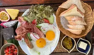 A top-down view of a hearty breakfast platter on a wooden table. The meal includes two fried eggs, bacon, sausage, a side salad, fresh tropical fruit like passionfruit and dragonfruit, and a basket of sliced bread and toast.