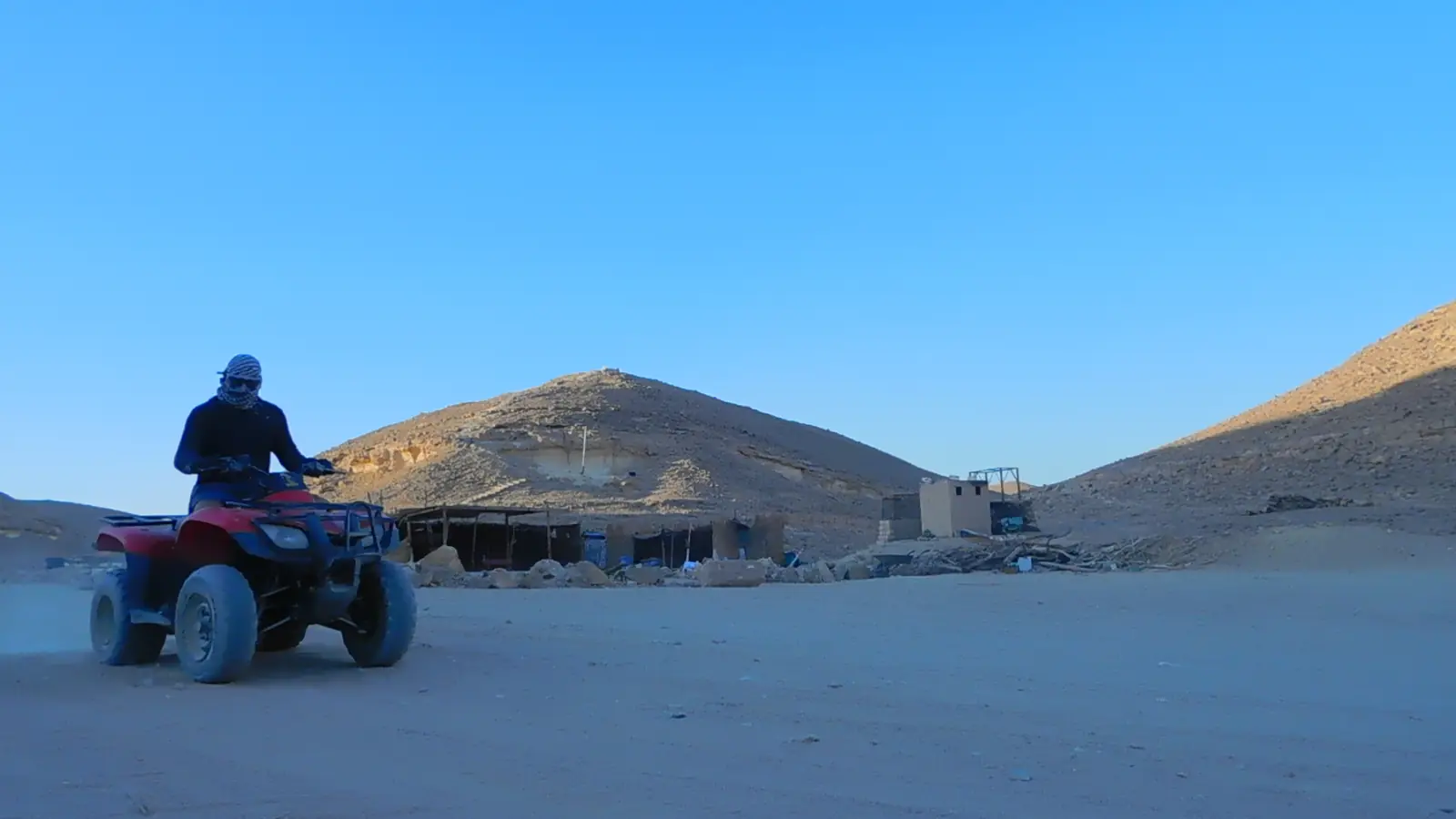 A person wearing a helmet and scarf rides a red ATV on a sandy desert terrain. The sky is clear, and the sun casts long shadows on the ground. Another person on an ATV can be seen in the background.