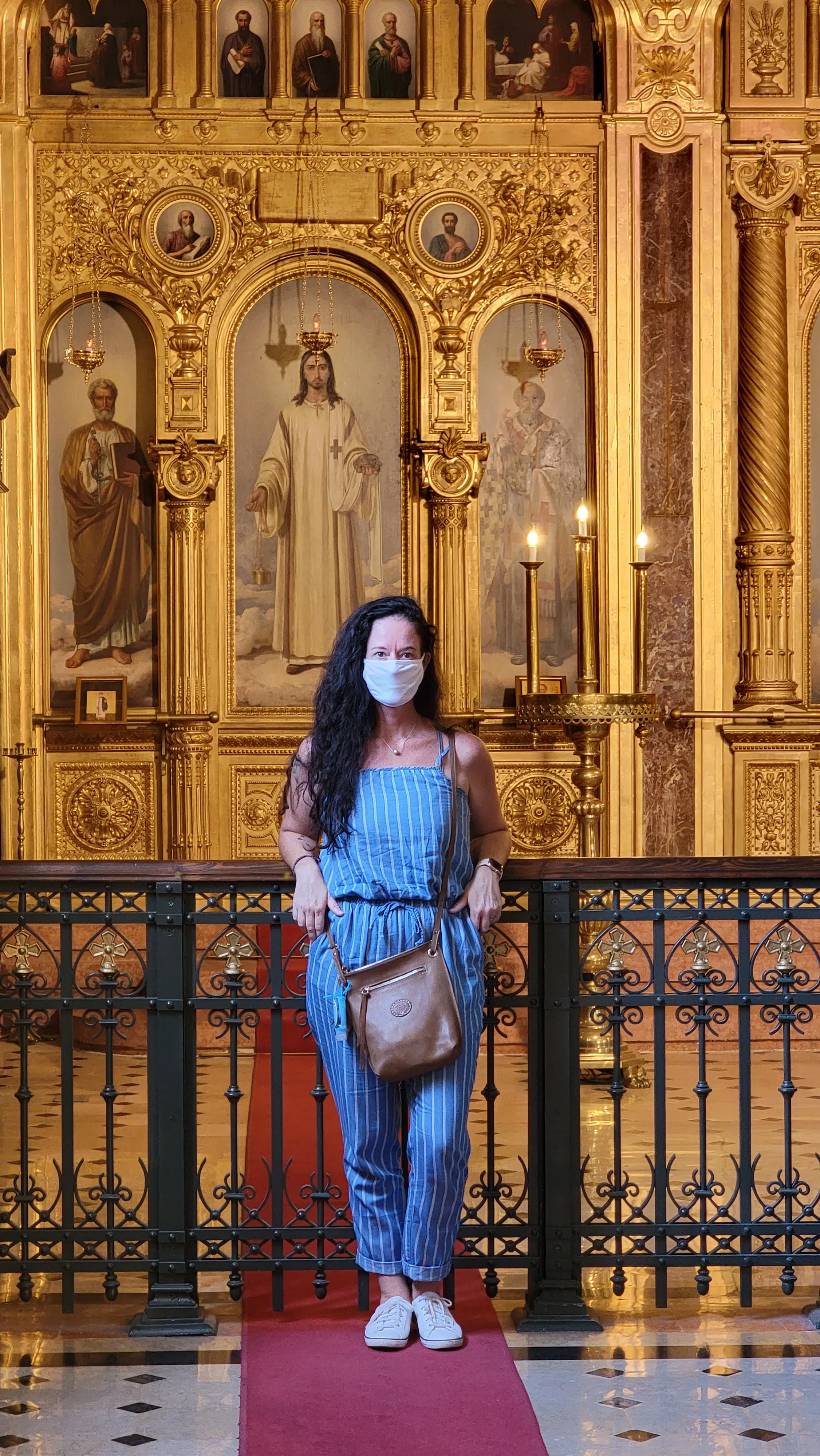 A woman with long dark hair stands on a red carpet in front of an ornate, golden religious altar, wearing a blue outfit and a face mask, with a brown crossbody bag.