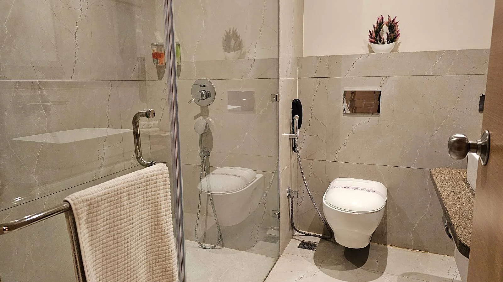 A modern bathroom with a glass-enclosed shower, a wall-mounted toilet, a bidet spray, a white towel on a rail, and a potted plant on a ledge above the toilet. The walls and floor are light-colored tiles.