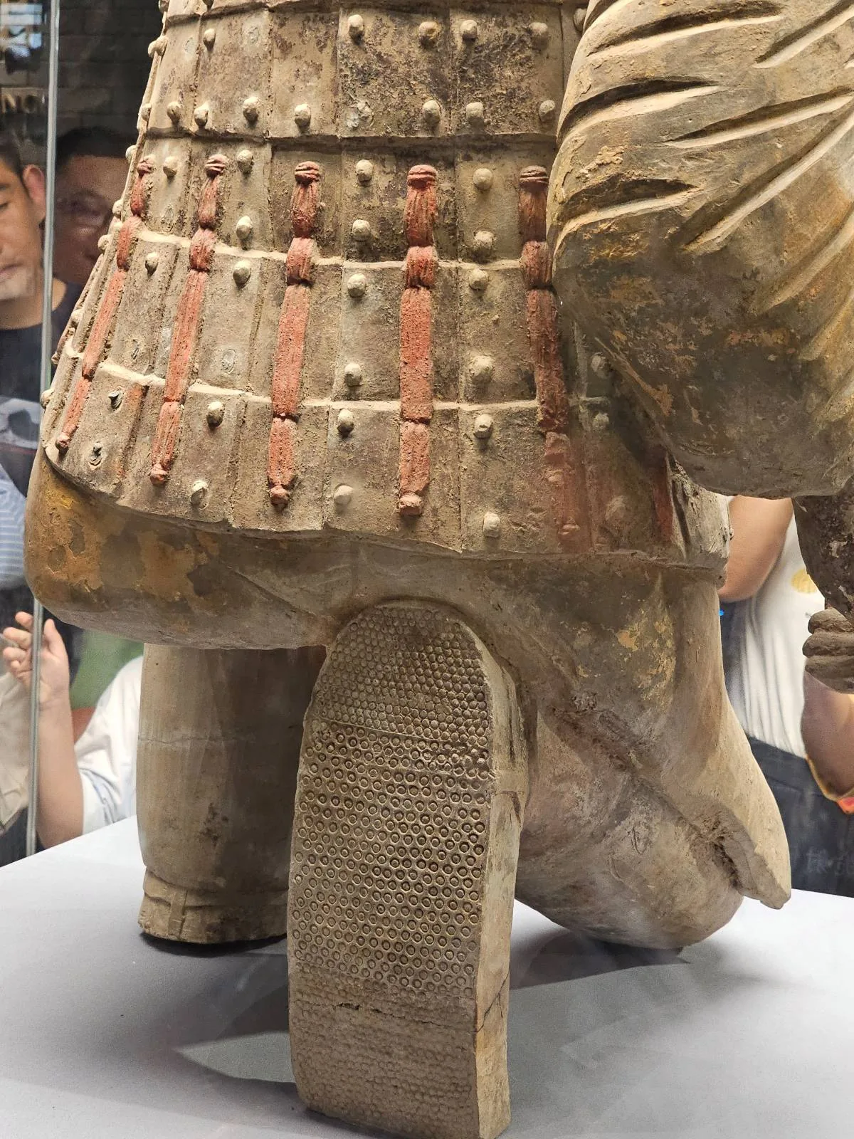 Close-up of the lower half of an ancient statue showing detailed armor and raised footwear, displayed in a museum with several people observing in the background.