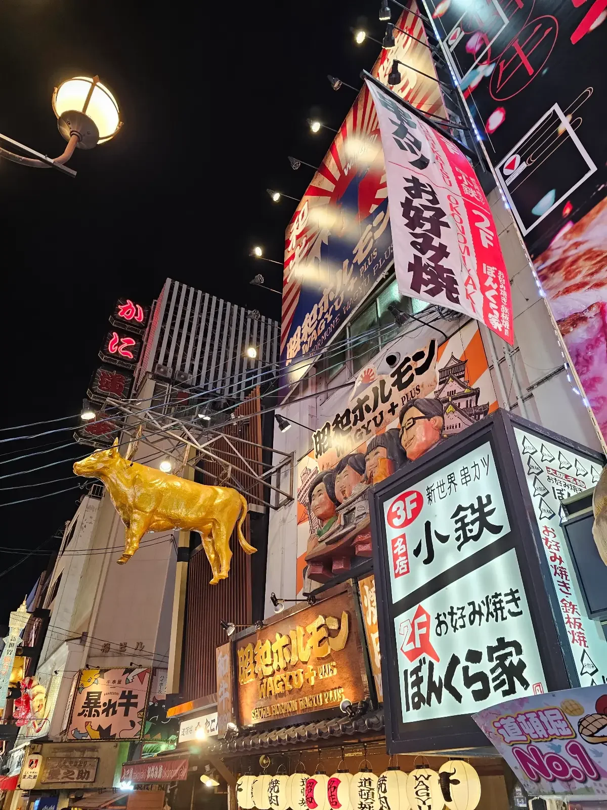 Brightly lit street at night with colorful signs in Japanese and a large golden cow statue hanging above the sidewalk, likely in a lively city area known for restaurants or entertainment.
