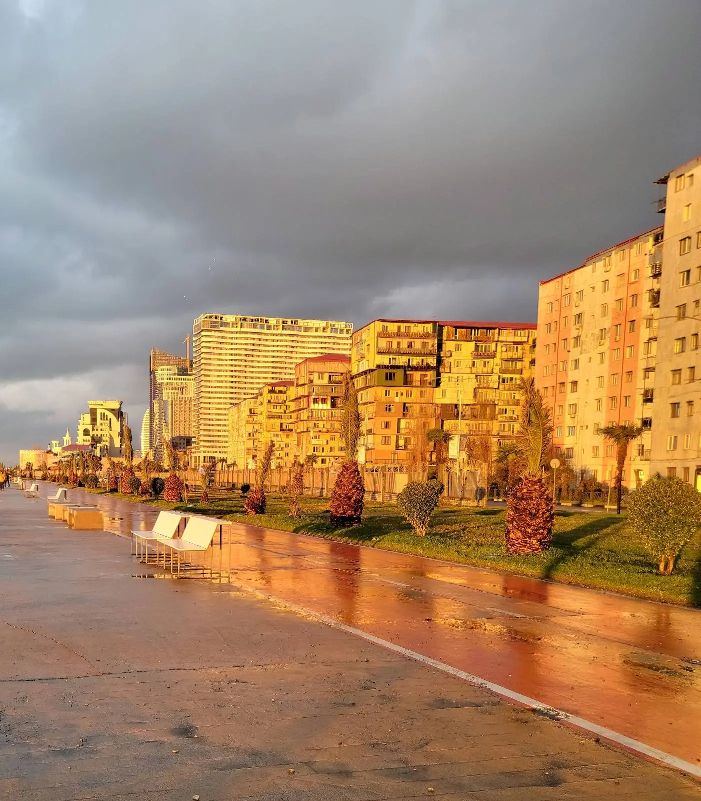 Buildings and a sidewalk are bathed in golden sunlight under a dramatic, cloudy sky; the wet pavement reflects the light, and there are trees and greenery along the street.