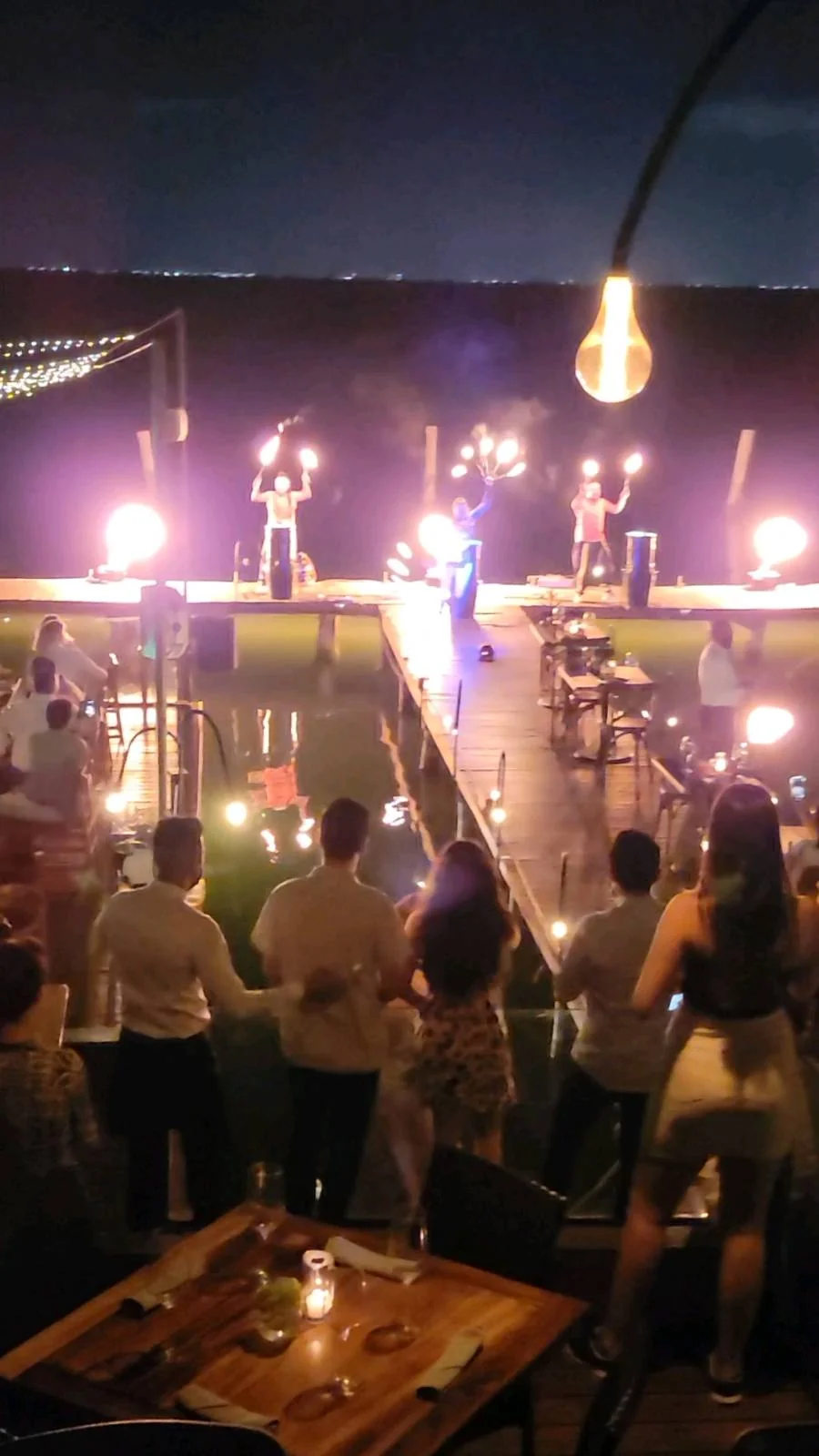A lively fire show by performers on a dock over water at night. Spectators watch, captivated by the flames. Tables with candles are in the foreground, creating a warm, festive atmosphere against the dark night sky.