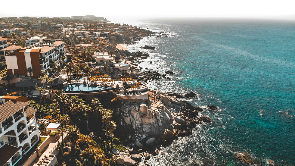 Aerial view of a coastal resort with pools and palm trees, adjacent to rocky shoreline and turquoise ocean. Sunny and serene atmosphere.