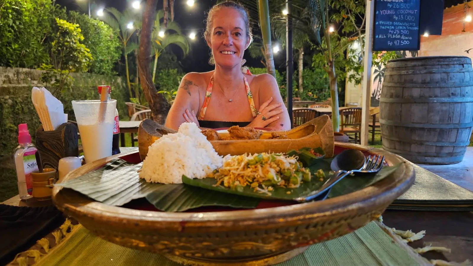 A person smiles at an outdoor table with various dishes in front, including rice and a vegetable stir-fry on a leaf. String lights and greenery are visible in the background, creating a cozy dining atmosphere.