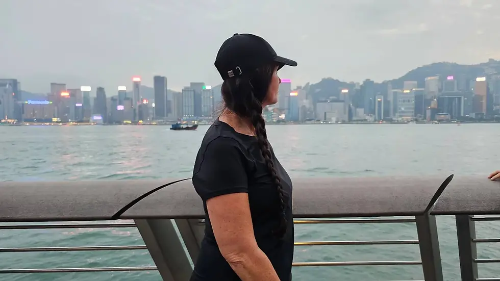 A person wearing a black cap and black shirt stands near a waterfront railing, with the Hong Kong city skyline and mountains in the background.