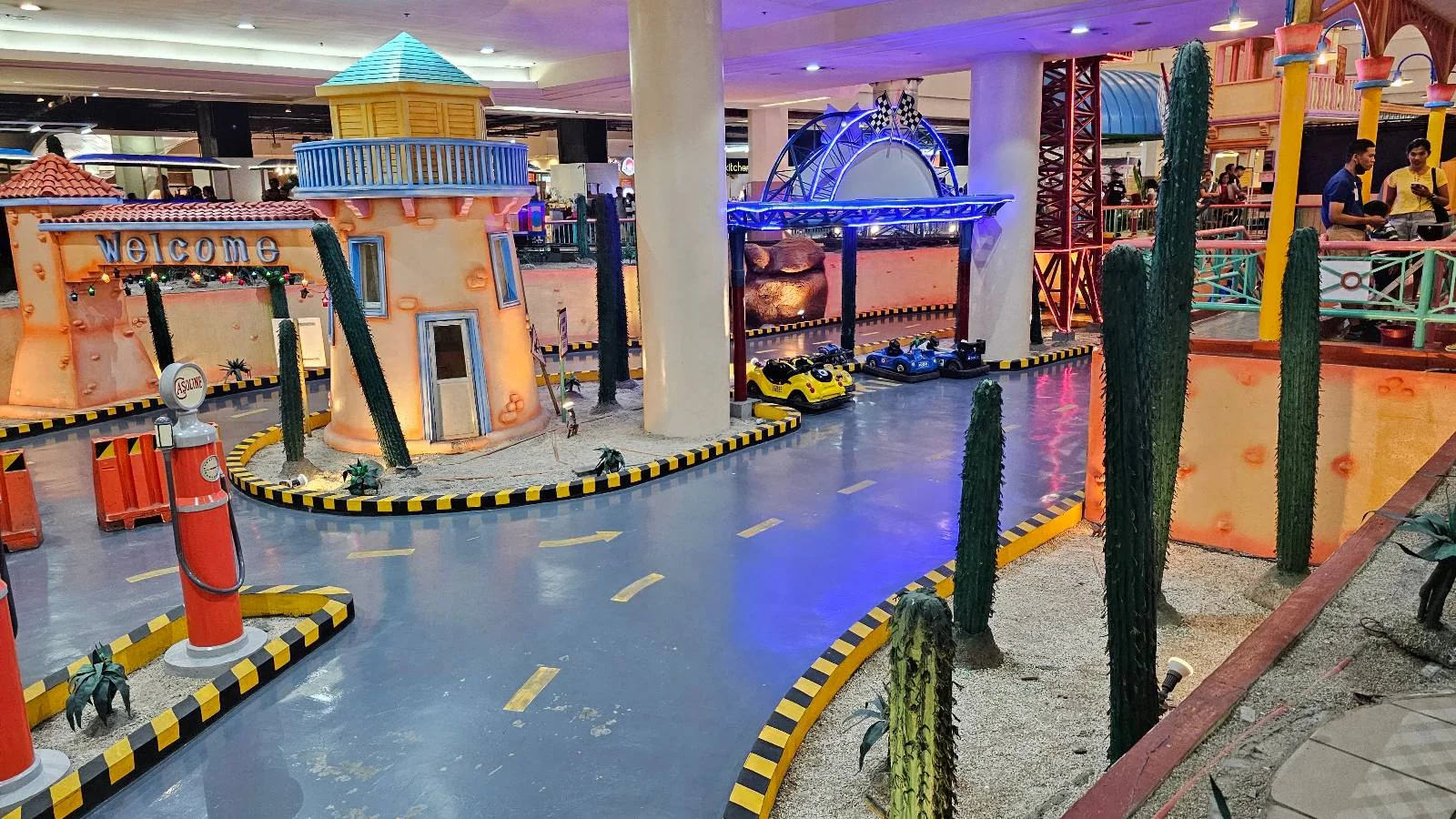Indoor amusement park track with colorful buildings, a winding path, and yellow and black safety markings. Cactus decorations are placed along the route. Bright lights illuminate the vibrant scene.