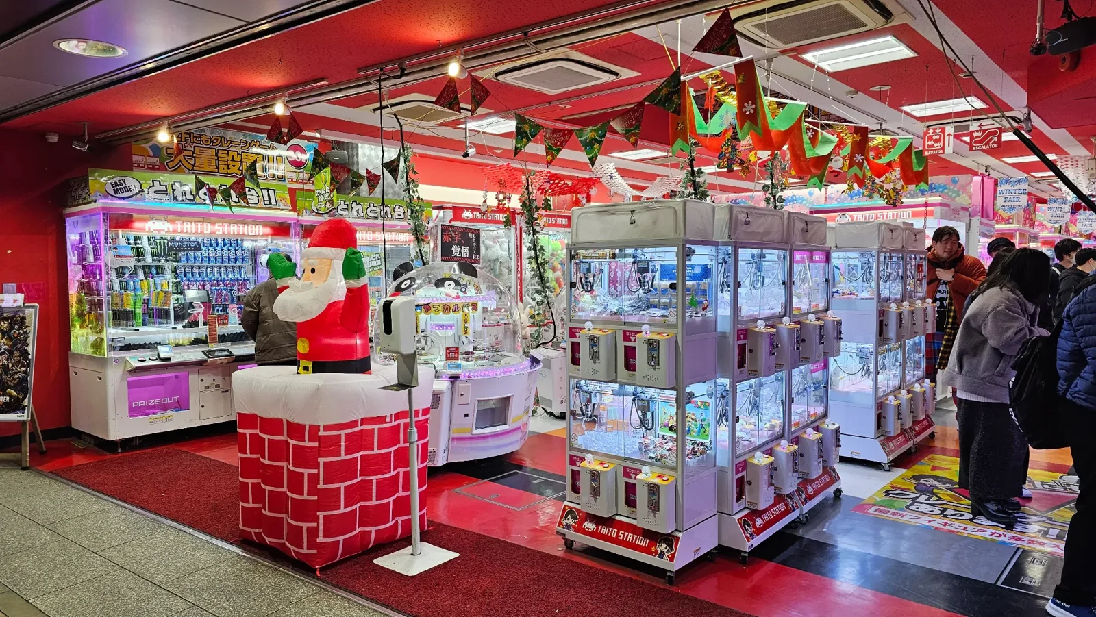 A brightly lit arcade filled with colorful claw machines and gashapon capsule toy dispensers, decorated with festive banners and a large Santa Claus figure at the entrance. Several people are playing inside.