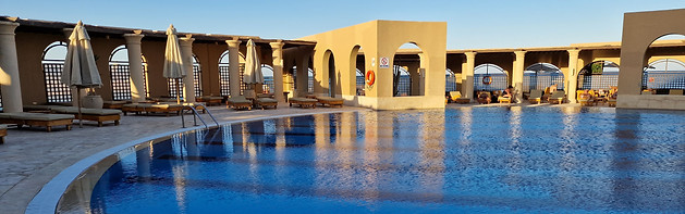 A serene outdoor swimming pool at a luxury resort is surrounded by beige stone columns and archways. The seating area includes sunbeds with umbrellas, and the blue sky is visible in the background. The pool reflects the structures, creating a peaceful atmosphere typical of boutique stays.