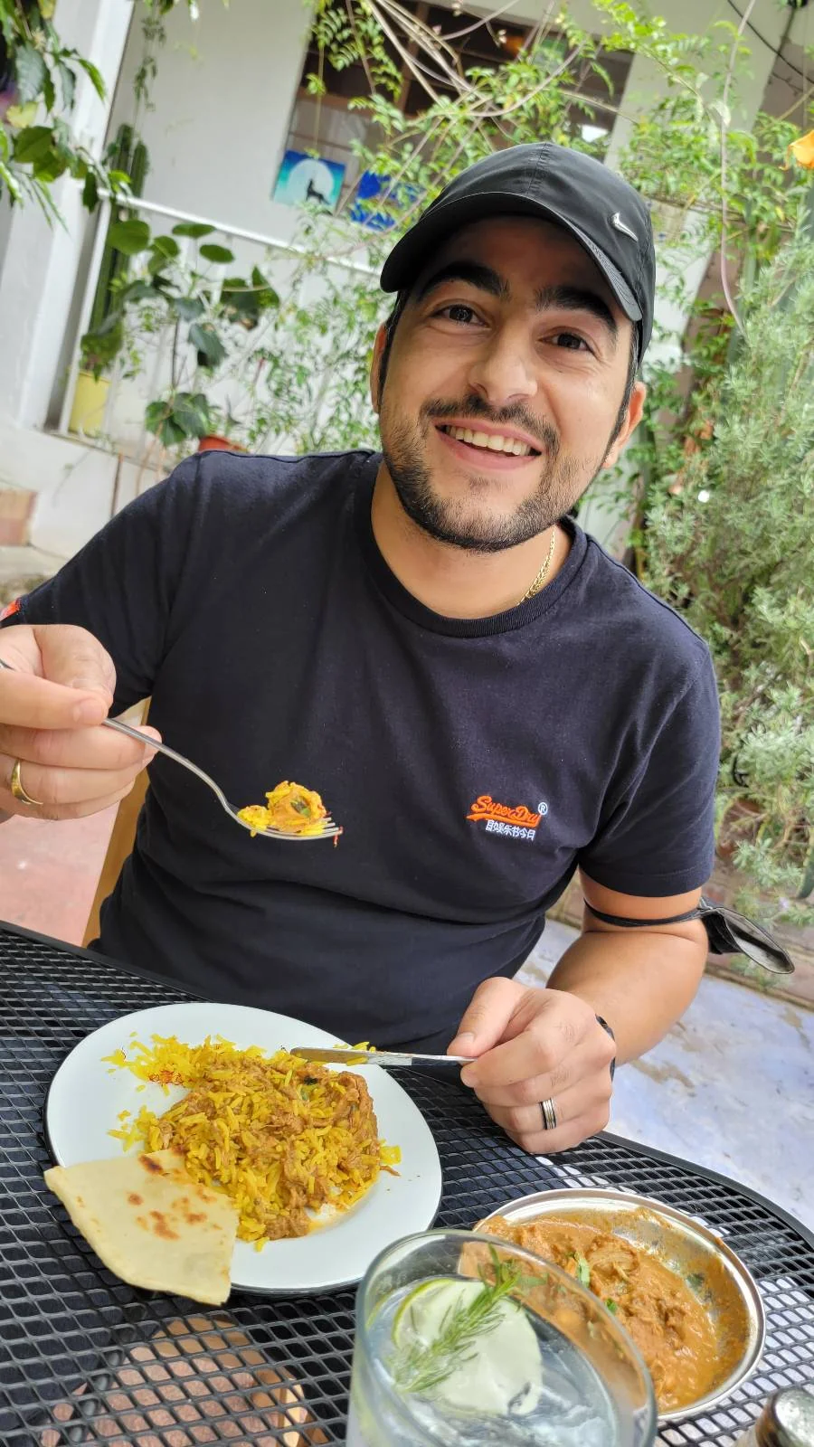A person wearing a cap and black t-shirt is sitting at an outdoor table. They are smiling and holding a fork over a plate of rice with a side of naan and curry. Greenery is visible in the background.