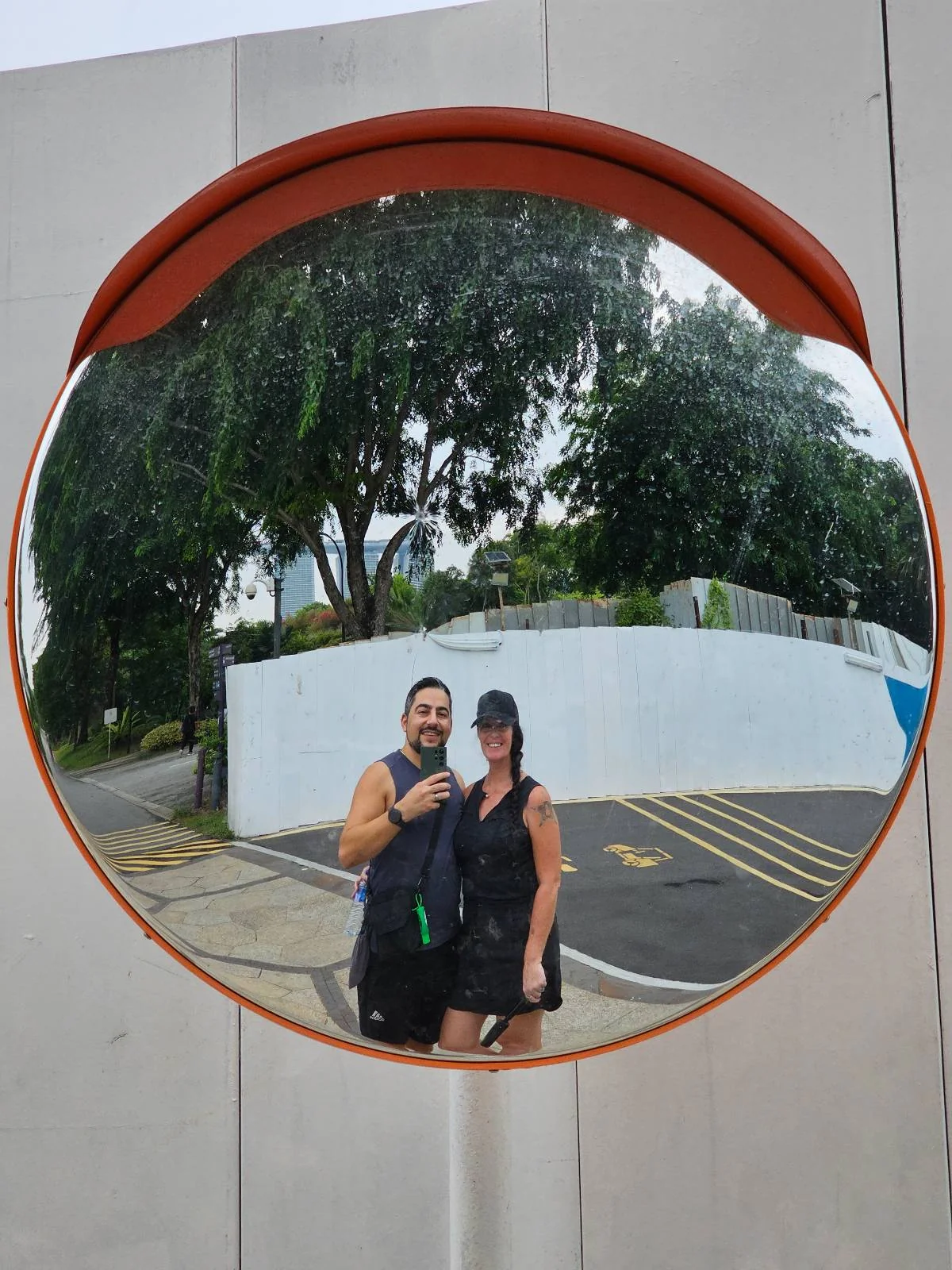 A man and woman are taking a selfie in a convex mirror on a street. They are smiling, and the mirror reflects surrounding greenery and road markings. The woman is wearing a hat, and they are both dressed casually.