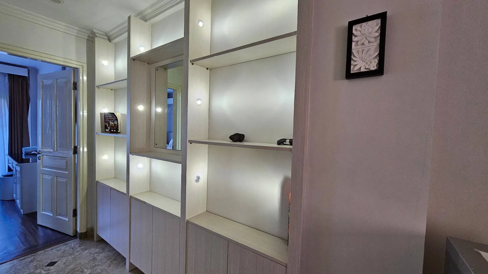 A hallway with built-in white illuminated shelves, some with small decorative items, next to a door leading to another room. A small framed artwork hangs on the adjacent pale wall.