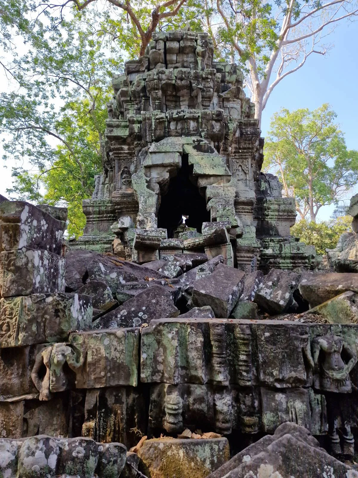 Ancient stone temple ruin with a large, jagged hole in the center, surrounded by fallen rocks and framed by green trees and a blue sky in the background.