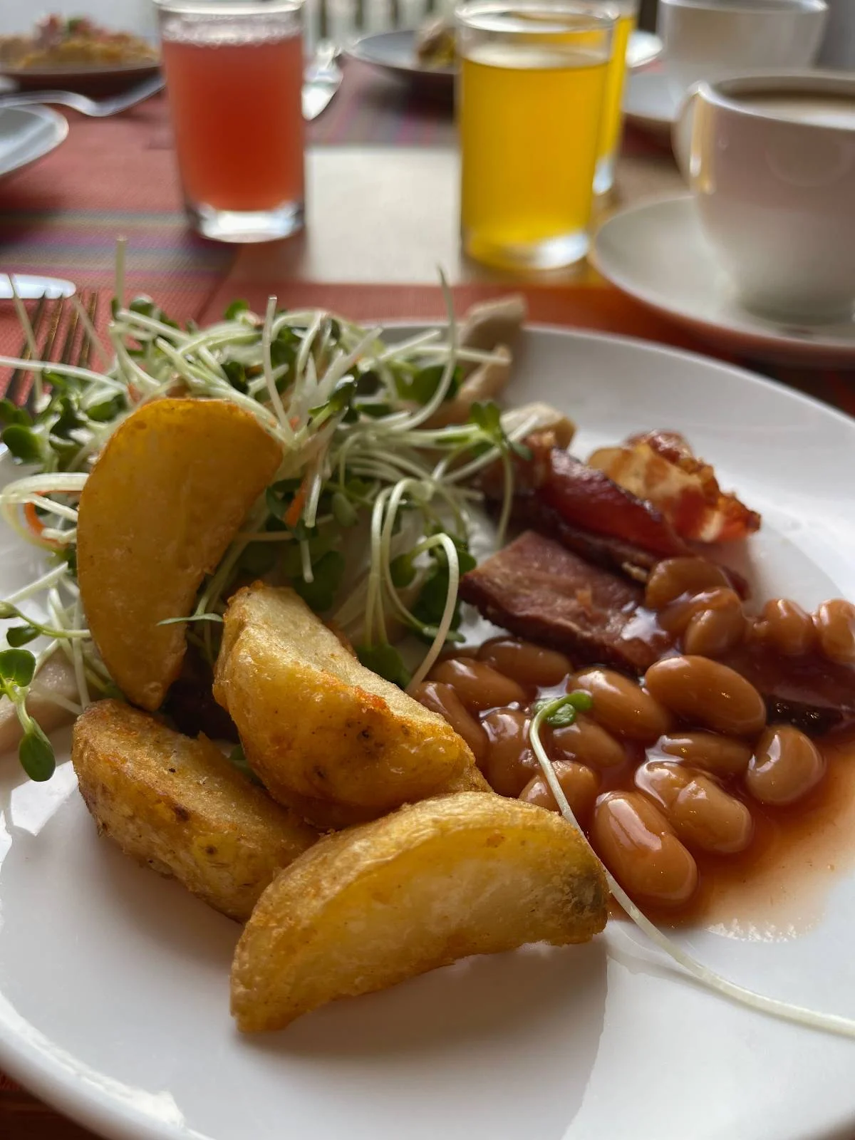 A plate of food featuring potato wedges, baked beans, greens, and slices of roasted meat with a lemon wedge. In the background, glasses with red and yellow beverages and a coffee cup, all on a wooden table.