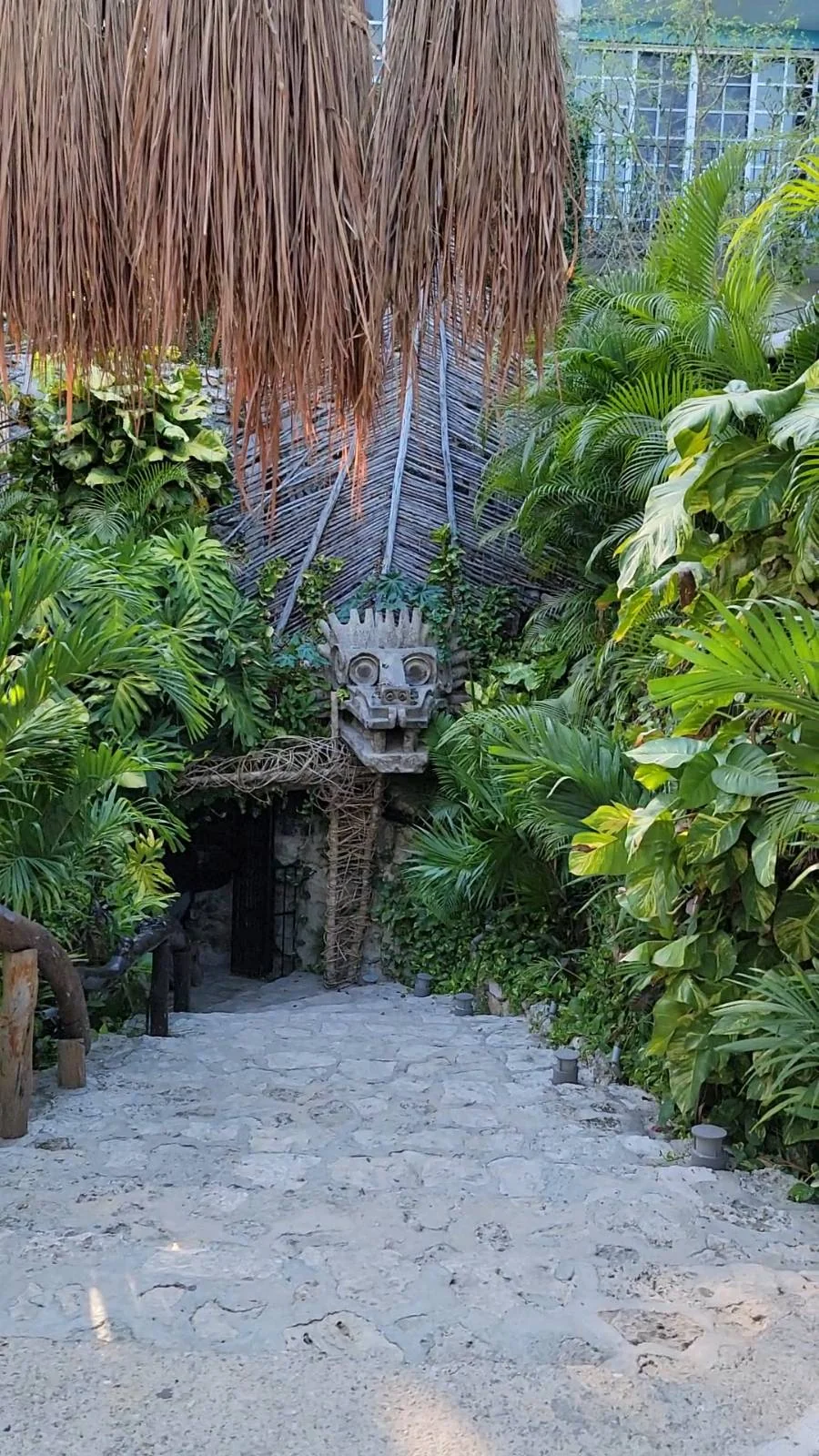 A stone pathway leads to a jungle-themed entrance adorned with lush green foliage. A carved stone face with an open mouth forms the doorway, surrounded by tropical plants and a thatched roof above.