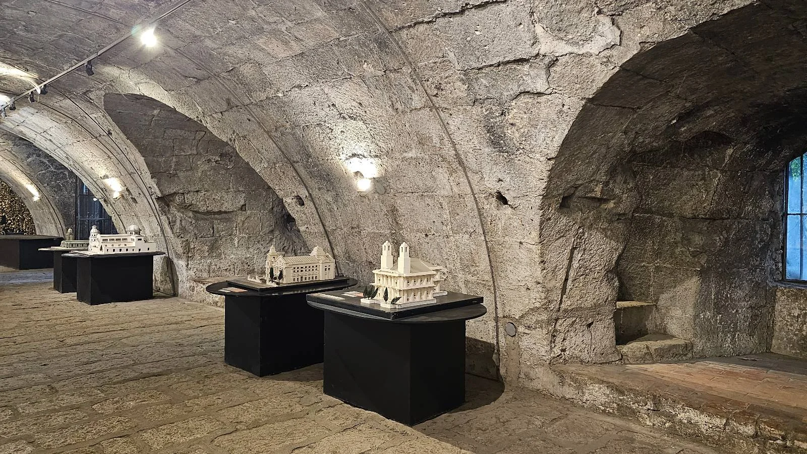 A dimly lit stone basement with arched ceilings. Several black tables display architectural models, illuminated by small overhead lights. The setting has a historic and rustic appearance.