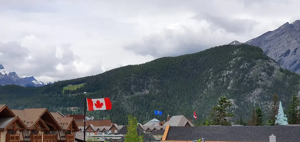 Canadian flag waving with mountains and cloudy sky in the background, surrounded by wooden buildings and trees.