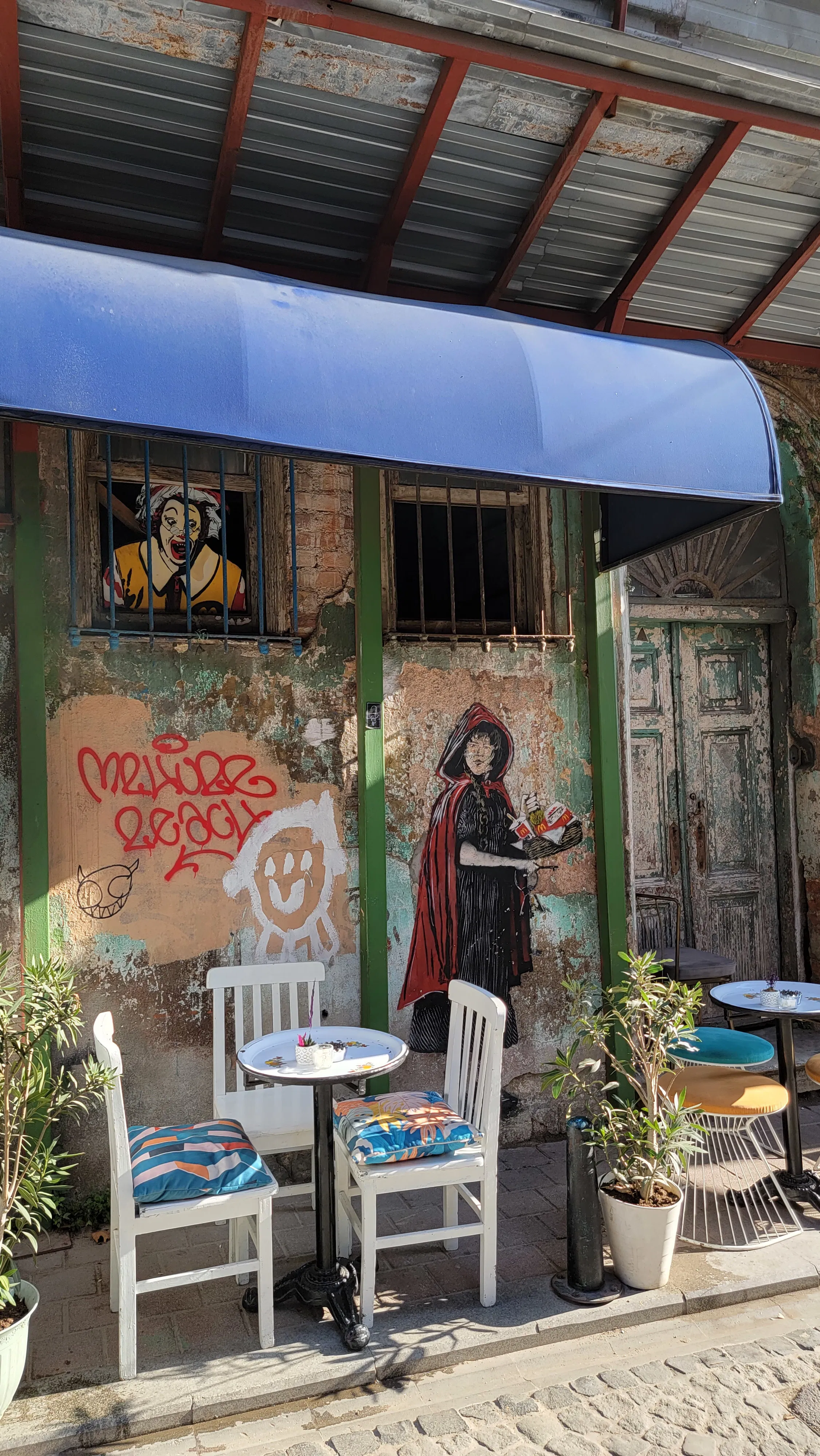 A small outdoor café with two white tables and blue chairs, potted plants, and colorful murals on the wall, including a woman holding a tray and a person in a yellow jacket, under a blue awning.