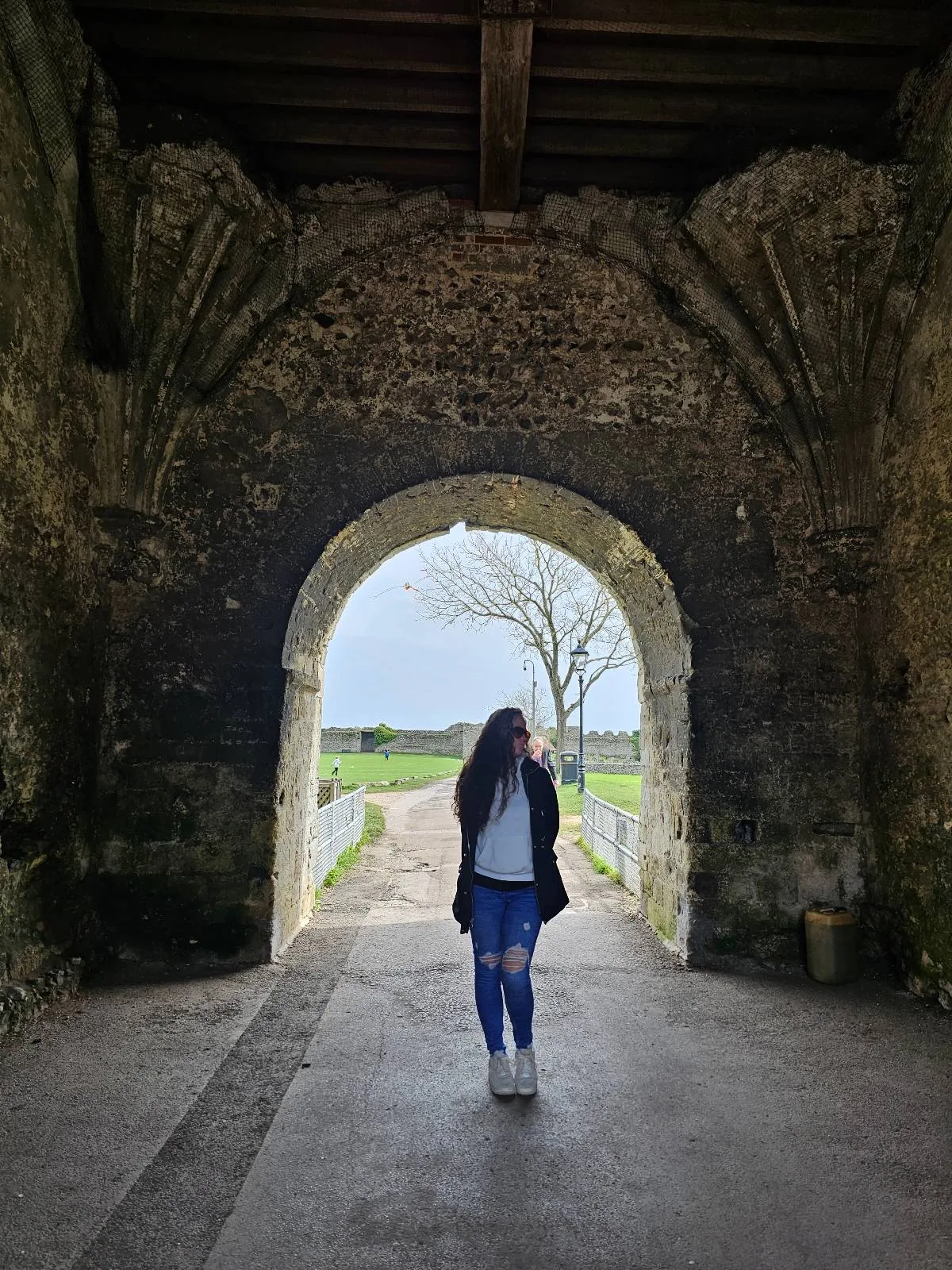 A person stands in the middle of a stone archway, looking to the side, with green fields and a leafless tree visible through the arch under a cloudy sky.