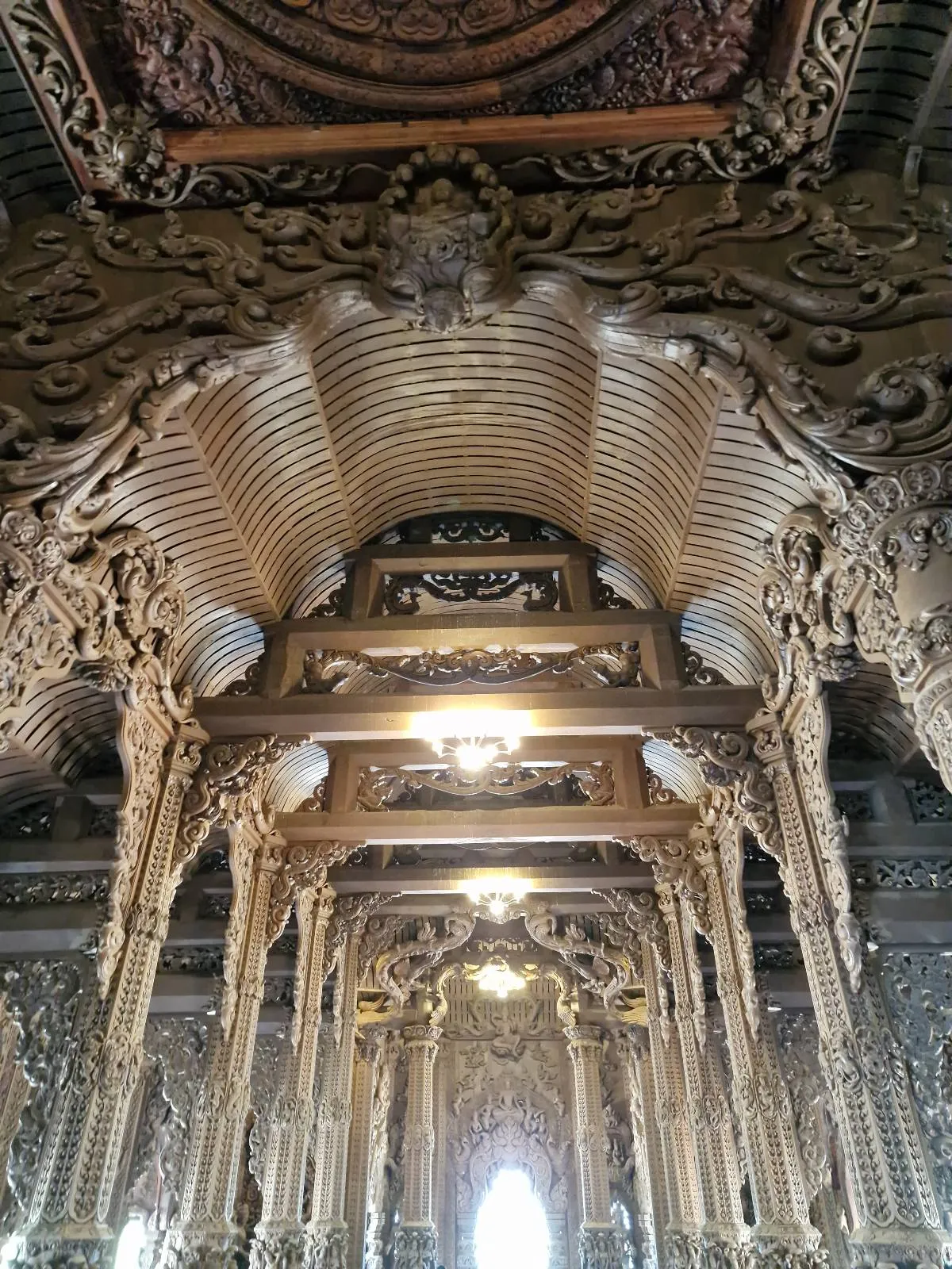 Intricately carved wooden ceiling with ornate decorations and arches, illuminated by soft lighting. The craftsmanship features detailed patterns and textures, creating an opulent and historical atmosphere.