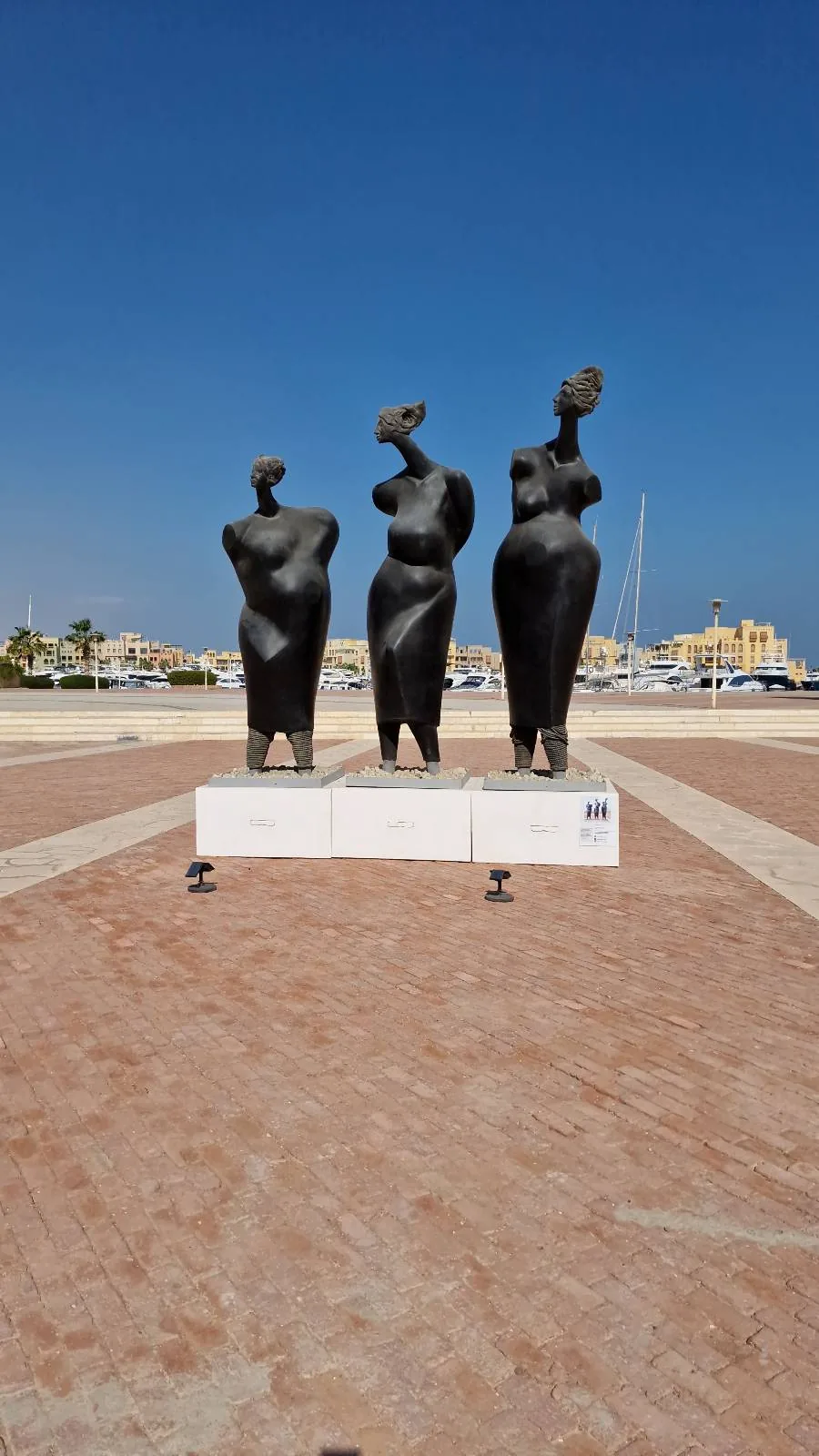 Three abstract female statues stand on a raised platform against a clear blue sky. The statues are dark and stylized, with simplified forms and rounded shapes.