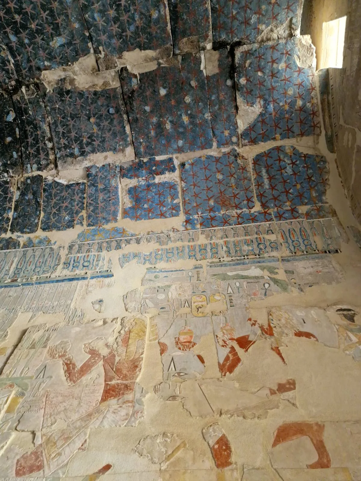 Interior of an ancient Egyptian tomb with faded wall paintings and inscriptions. The upper walls are decorated with blue tiles featuring red dots, while the lower section has deteriorated art and hieroglyphs. Natural light enters from the right.