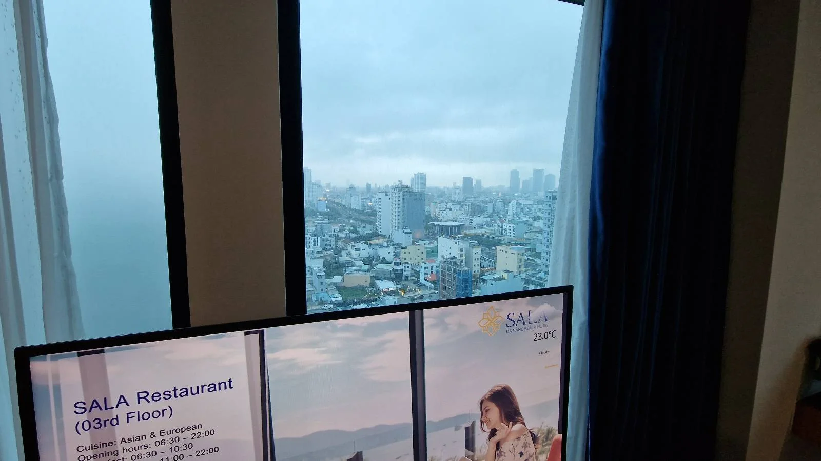 A hotel room with a large window showcasing a city skyline under a cloudy sky. A TV is visible, displaying an advertisement for a restaurant, with an image of a woman and a scenic view. Navy curtains frame the window.